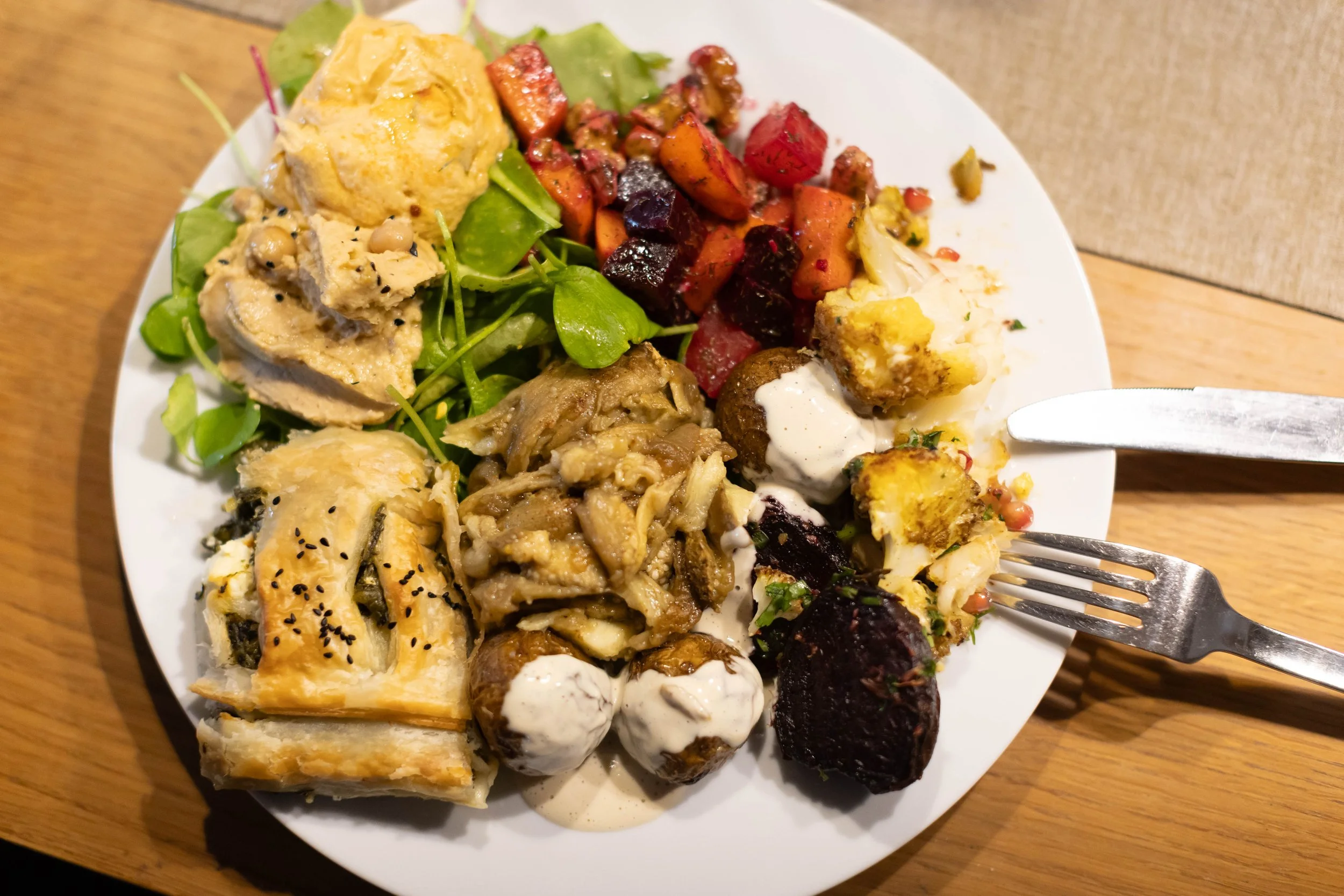A white plate of assorted foods including mixed vegetables, salad greens, a slice of vegetarian pie, roasted mushrooms, boiled potatoes with sauce, and beet slices, on a wooden table.