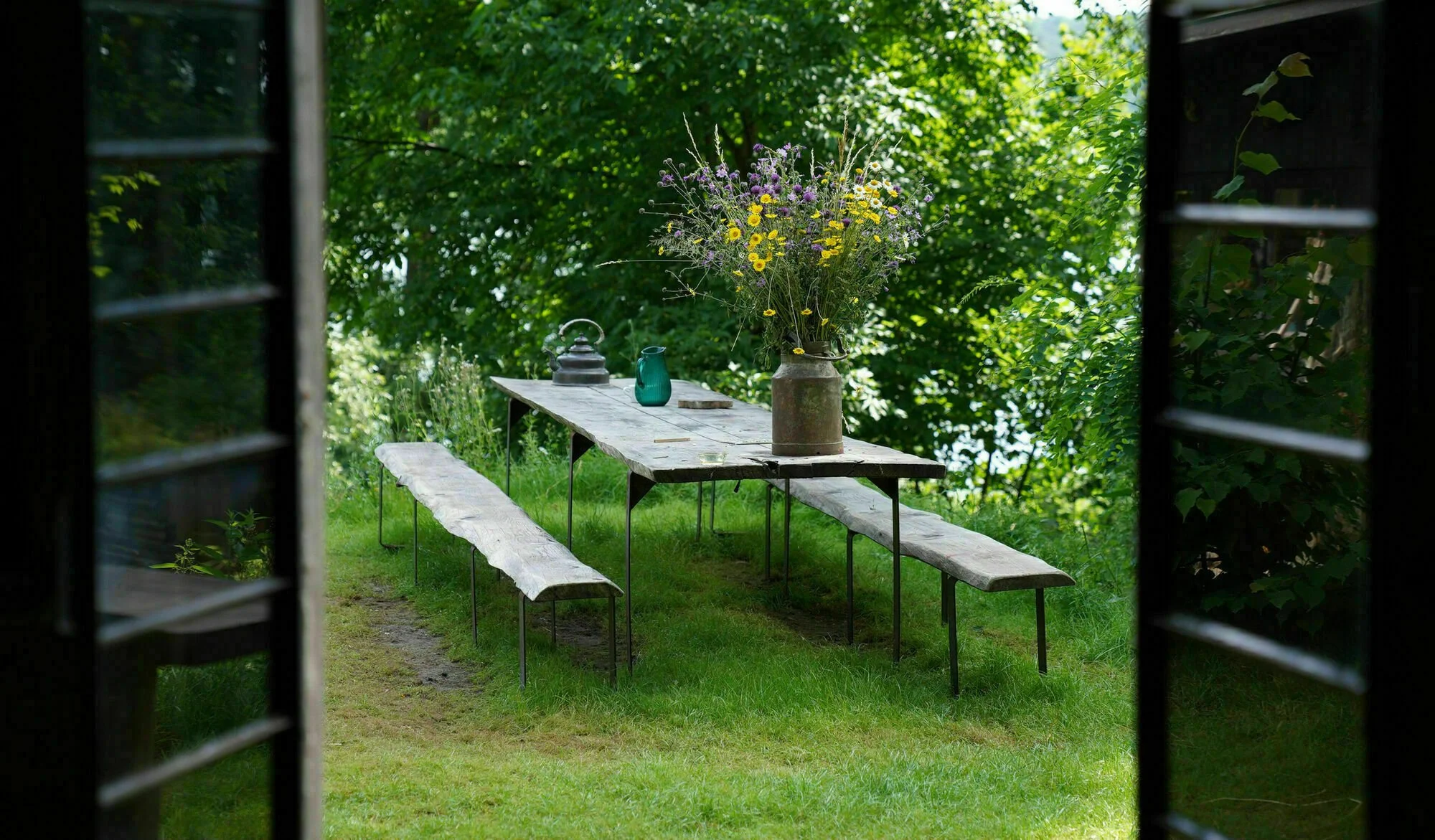 A rustic outdoor table with wooden benches on grassy ground, viewed through open window shutters. The table has a large vase with wildflowers, a teal kettle, and a small tray.