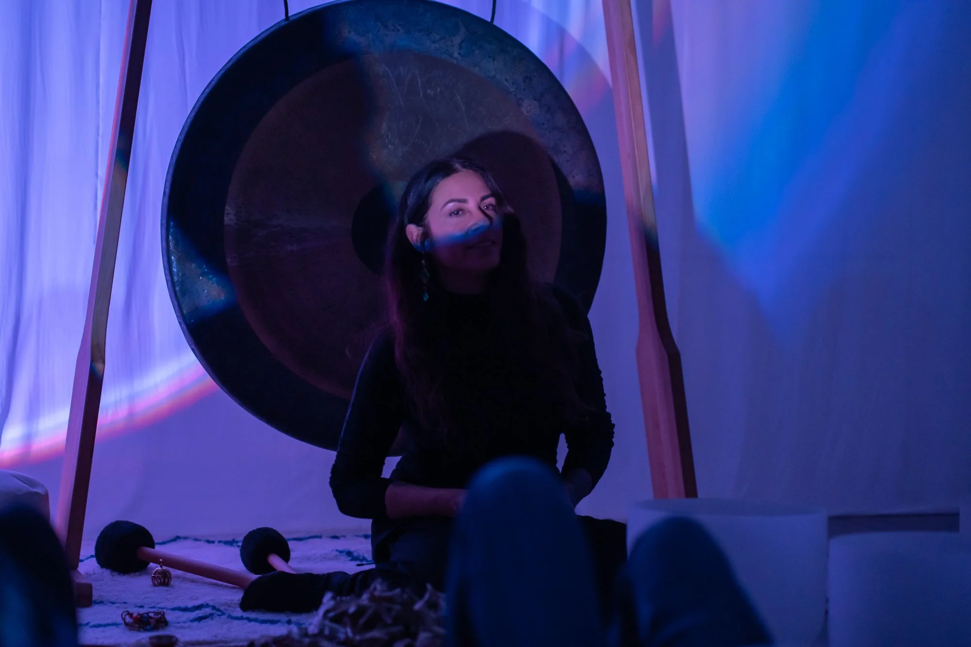 A woman with long dark hair sitting on the floor in front of a large gong, illuminated by purple and blue lighting, with small objects and a mallet nearby.