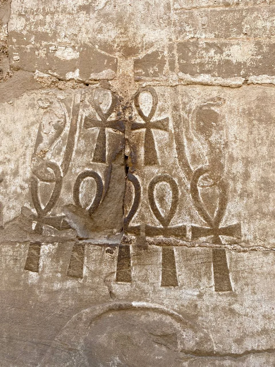 Ancient stone carving of three Ankh symbols with a cross pattern below, etched into a weathered wall.