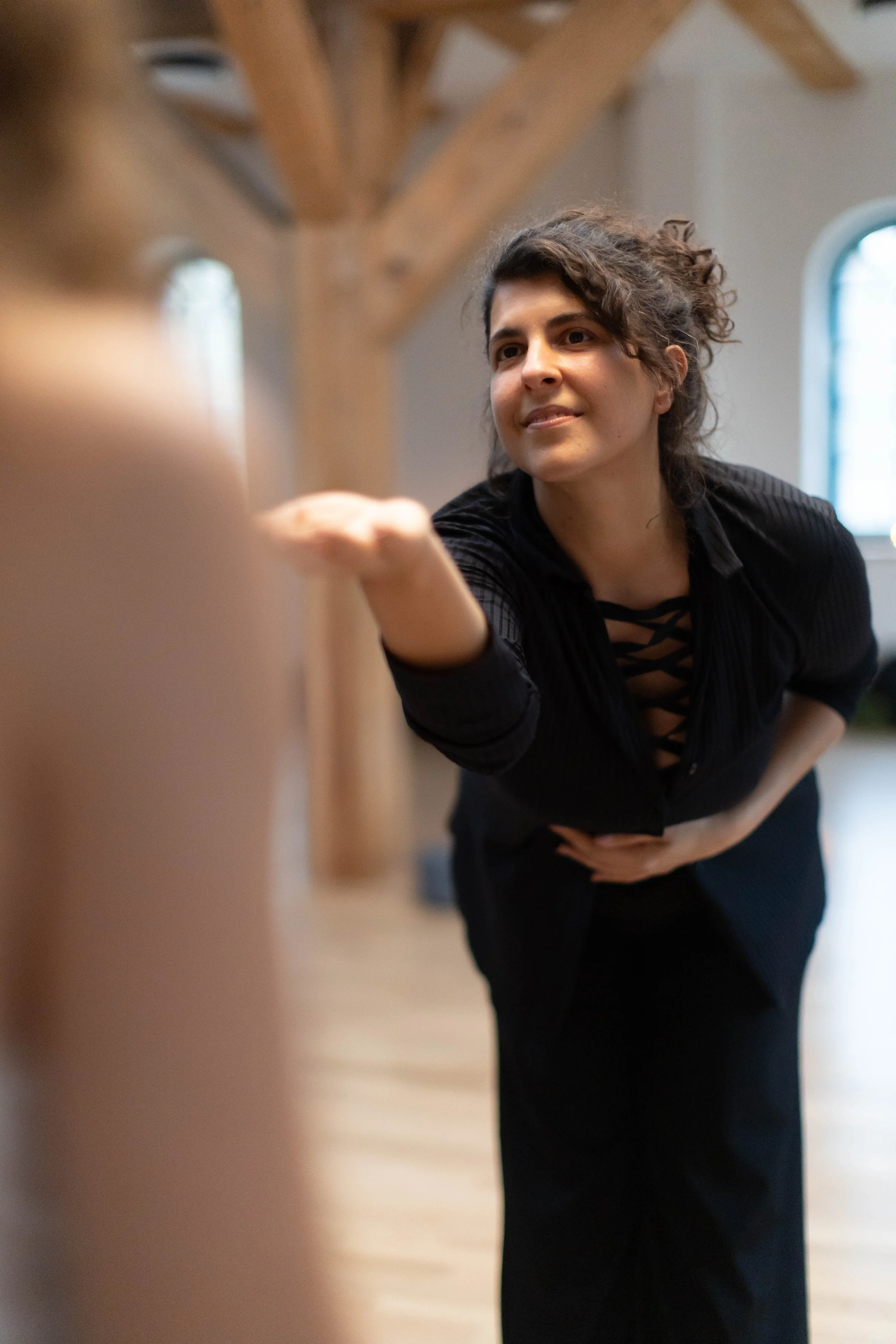 A woman with curly hair wearing a black top gestures with her hand while holding her stomach, engaging in a conversation or giving instructions inside a room with wooden beams and a window in the background.