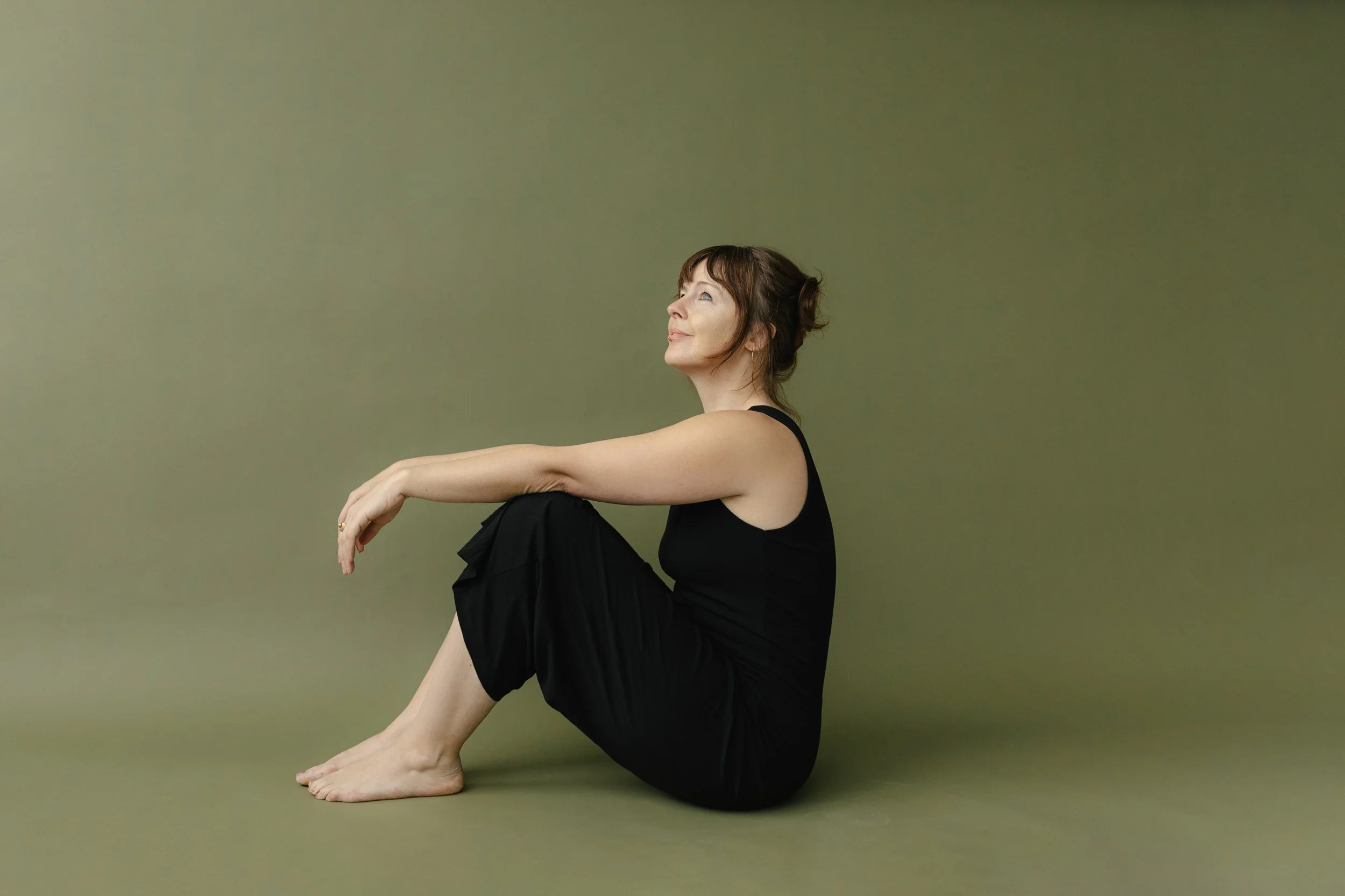 A woman sitting on the floor with her knees drawn up, arms resting on her knees, smiling and looking upward against a plain green background.