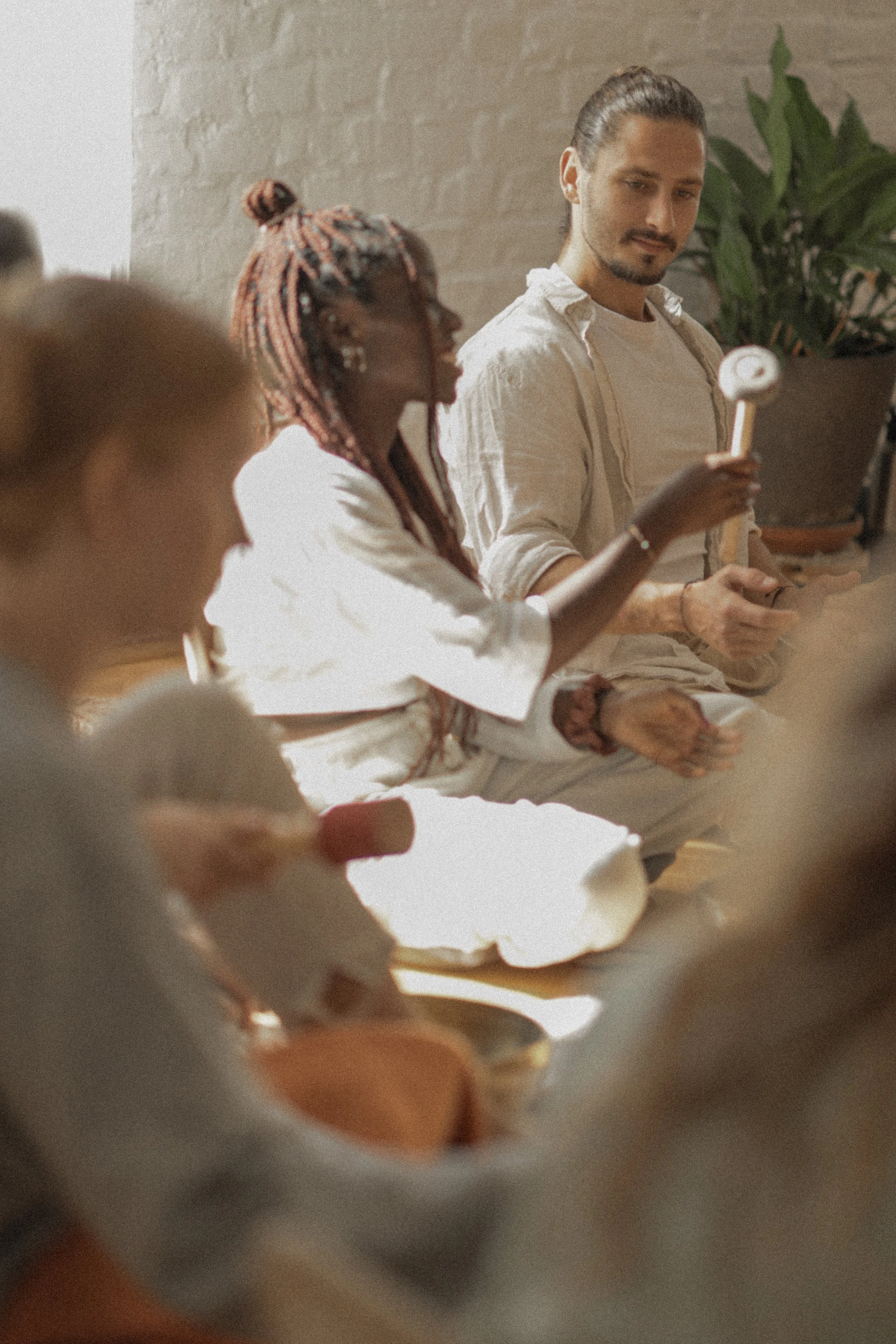 People sitting together indoors, one woman with colorful braids holding a wooden mallet, and a man with a beard and long hair tied back, looking at her, with others partially visible in the foreground.