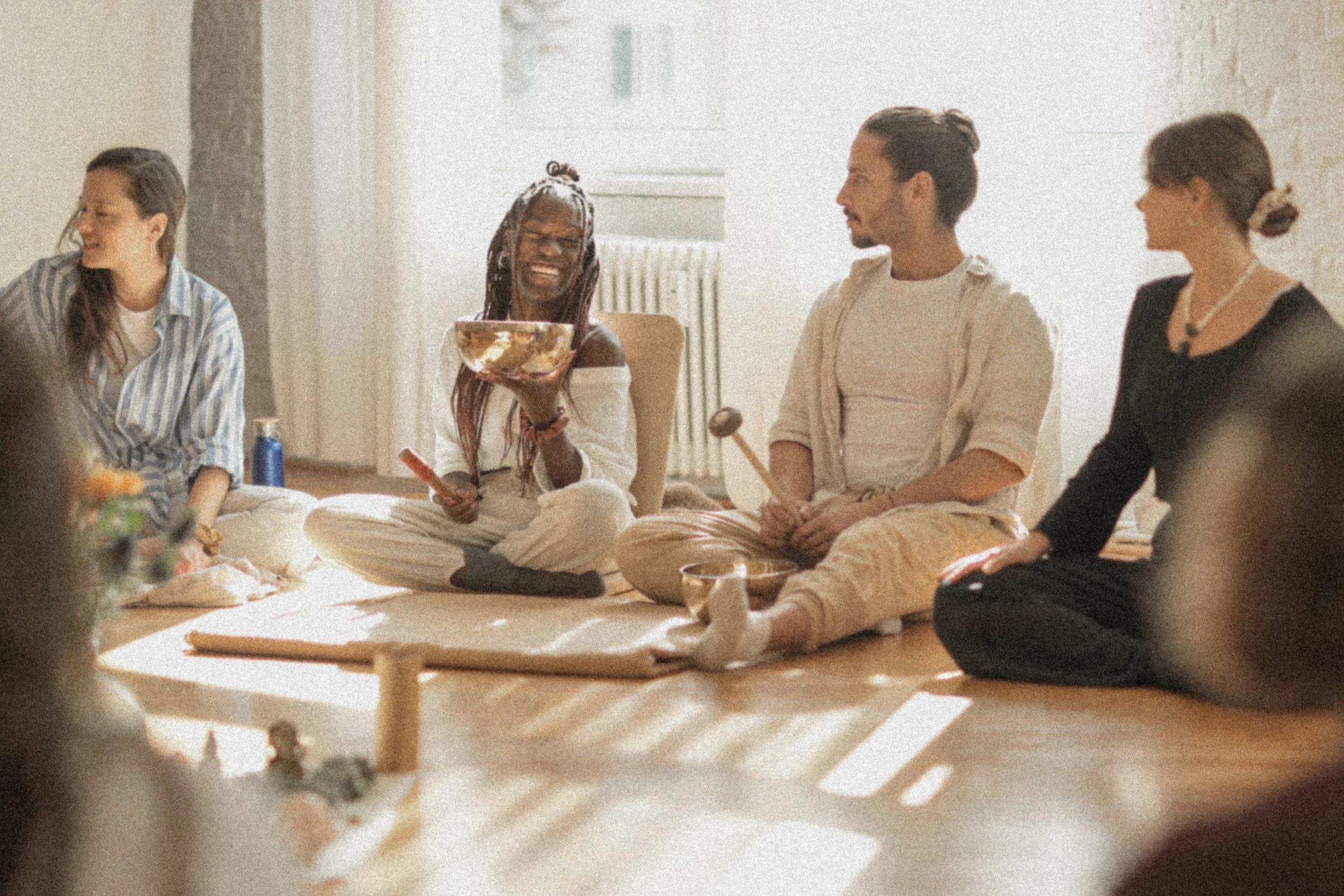Group of friends sitting on the floor, smiling and enjoying a gathering in a bright room, with one person holding a bowl.