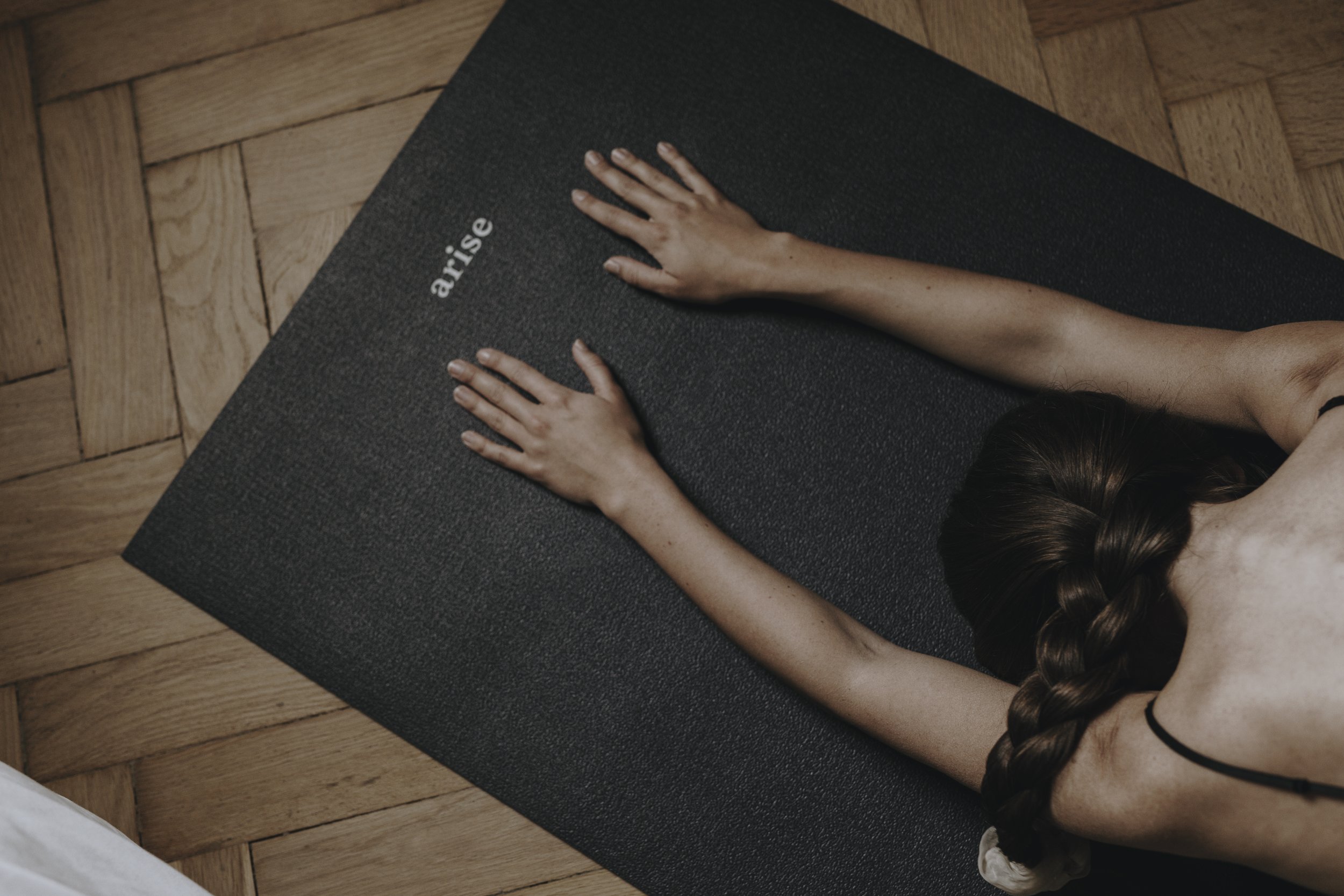 A woman with braided hair practicing yoga on a black mat labeled 'arise' on a wooden floor.