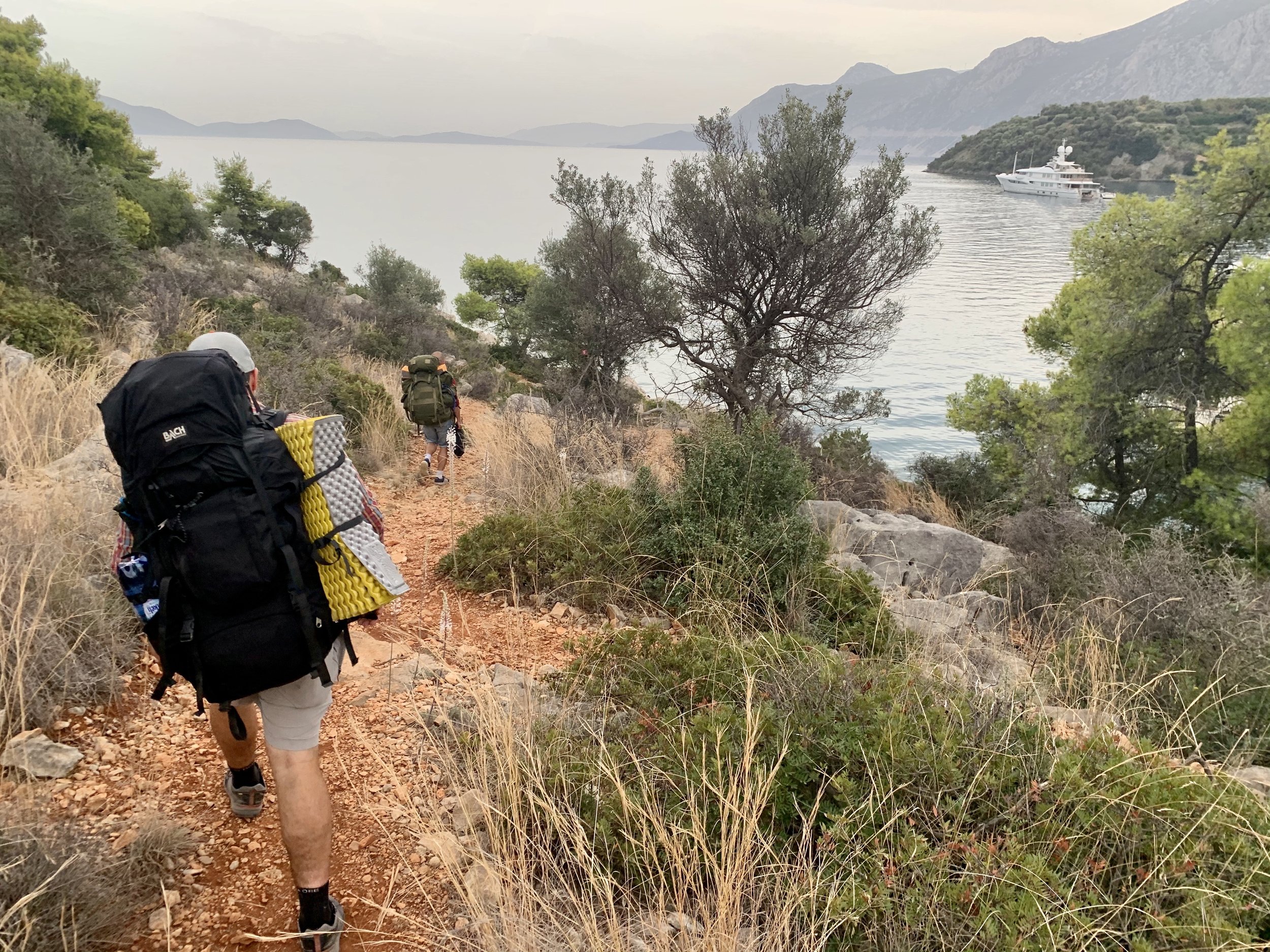 Three hikers walking along a dirt trail by a body of water with boat, surrounded by trees and mountains.