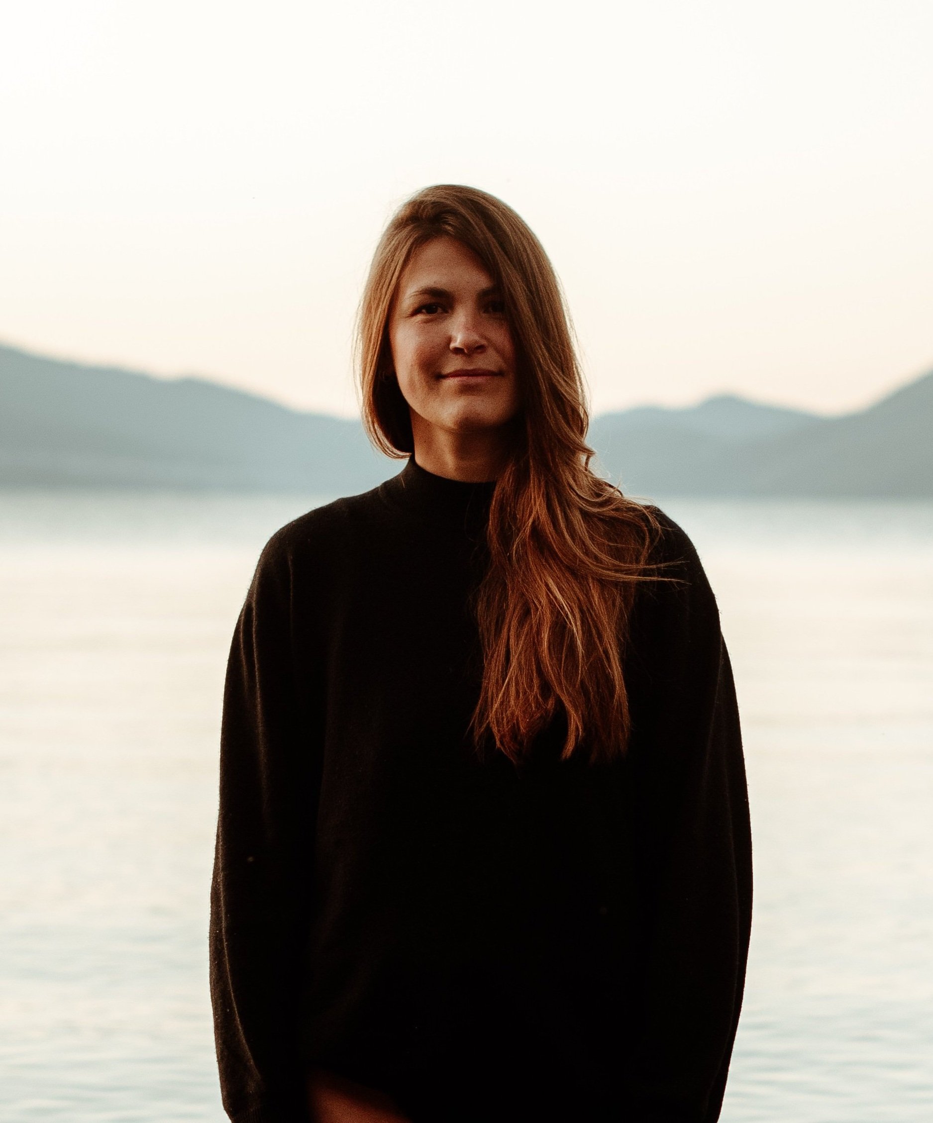 A woman with long reddish-brown hair wearing a black top standing by a lake with mountains in the background during sunset.