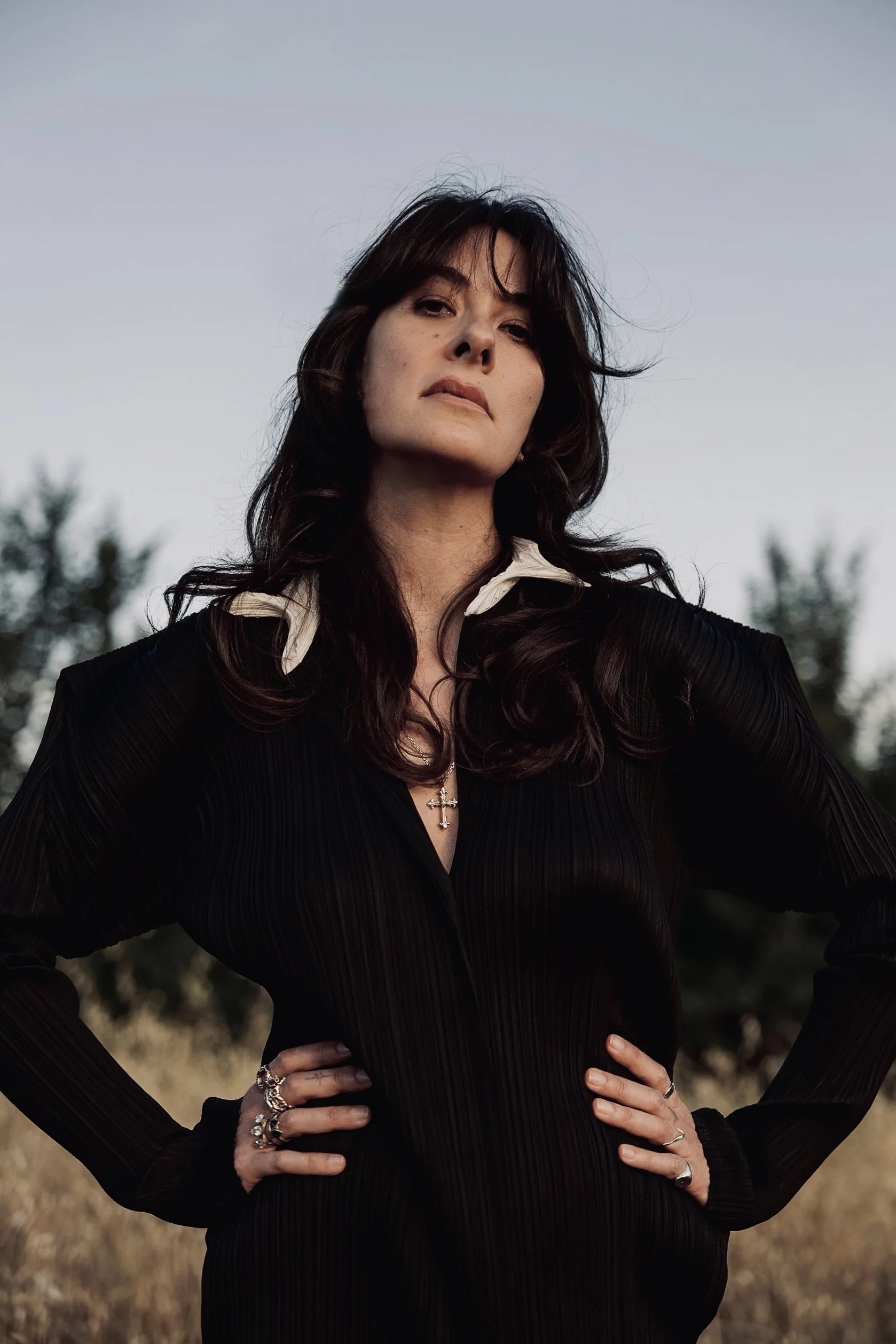 Woman with dark wavy hair standing outdoors with hands on hips, wearing a black pleated dress, white collar, and jewelry, against a background of trees and a clear sky.