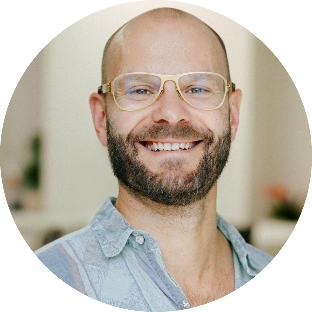 Smiling man with glasses and a beard, wearing a light-colored button-up shirt, indoors.
