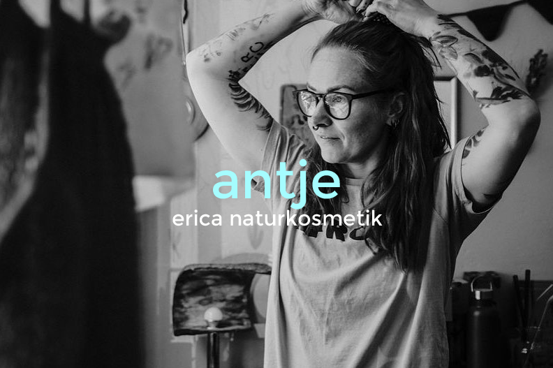 Black and white photo of a woman with glasses and tattoos, smiling with her hair held up by her hands, in a room with artwork on the walls.