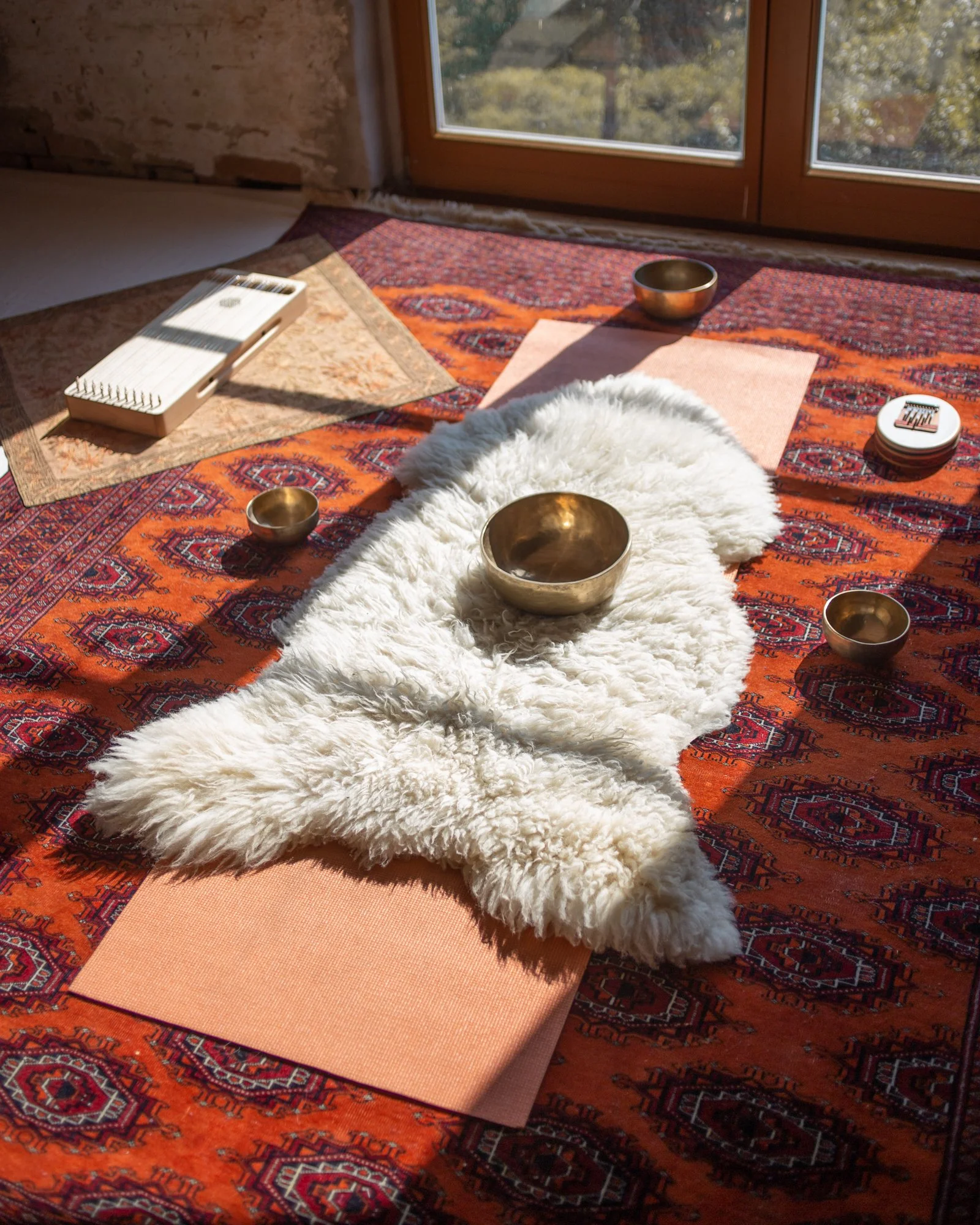 Yoga or meditation setup with brass singing bowls, a white sheepskin, pink yoga mats, an electronic musical instrument, and a box of singing bowls on a colorful patterned rug near a window.
