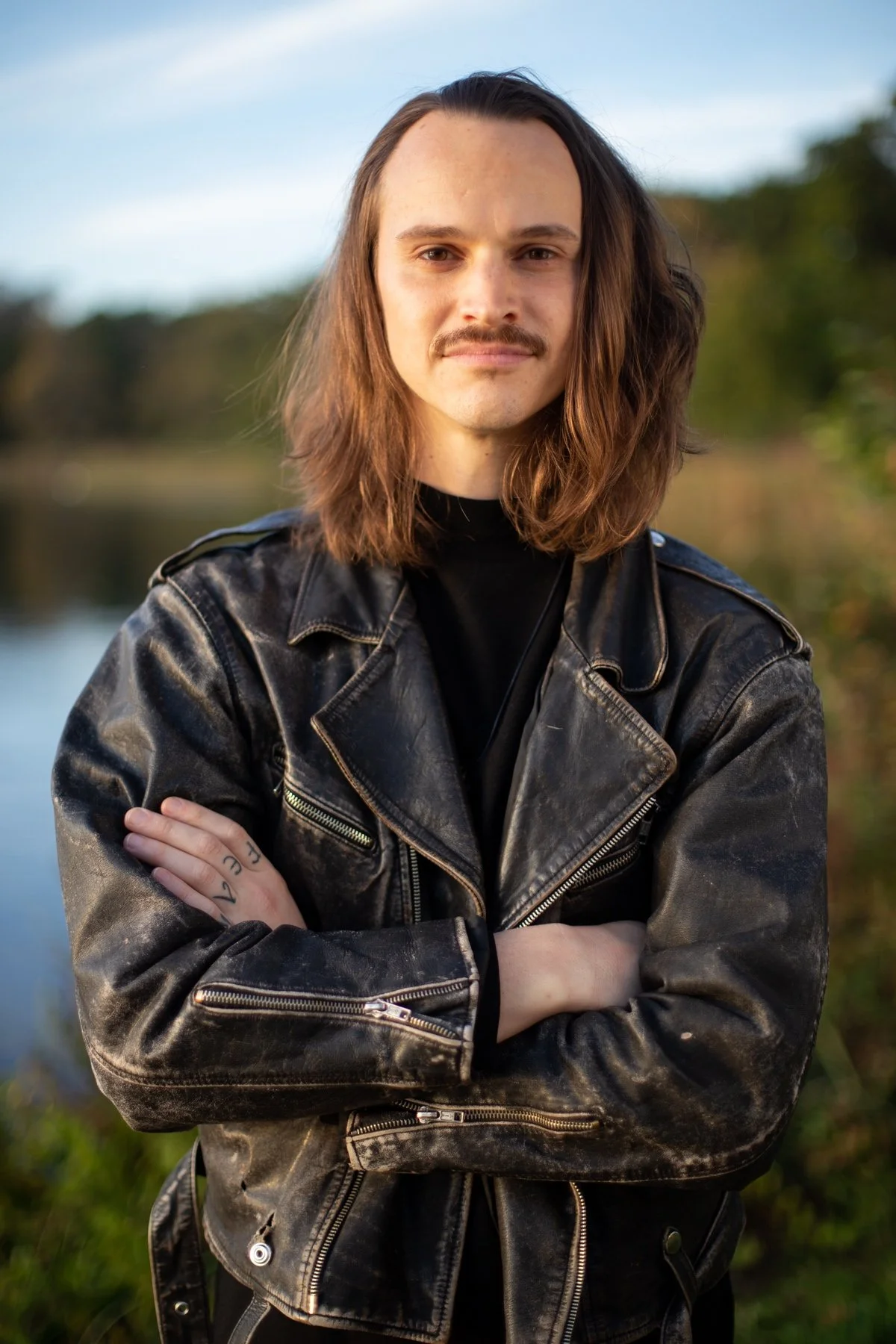 A person with long brown hair and a mustache, wearing a black leather jacket, standing outdoors near a body of water with trees in the background.