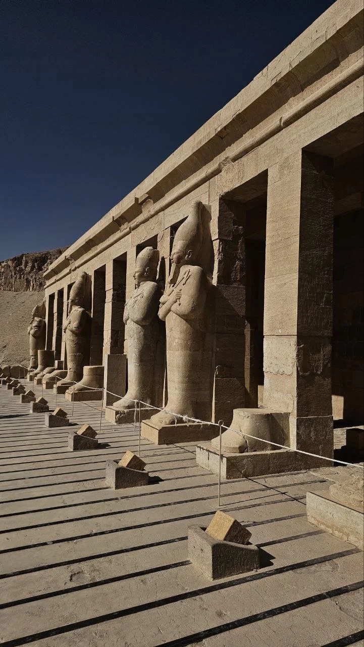 Ancient Egyptian statues of Pharaohs and deities standing in a row at an archaeological site, with a stone structure behind them and a clear sky above.