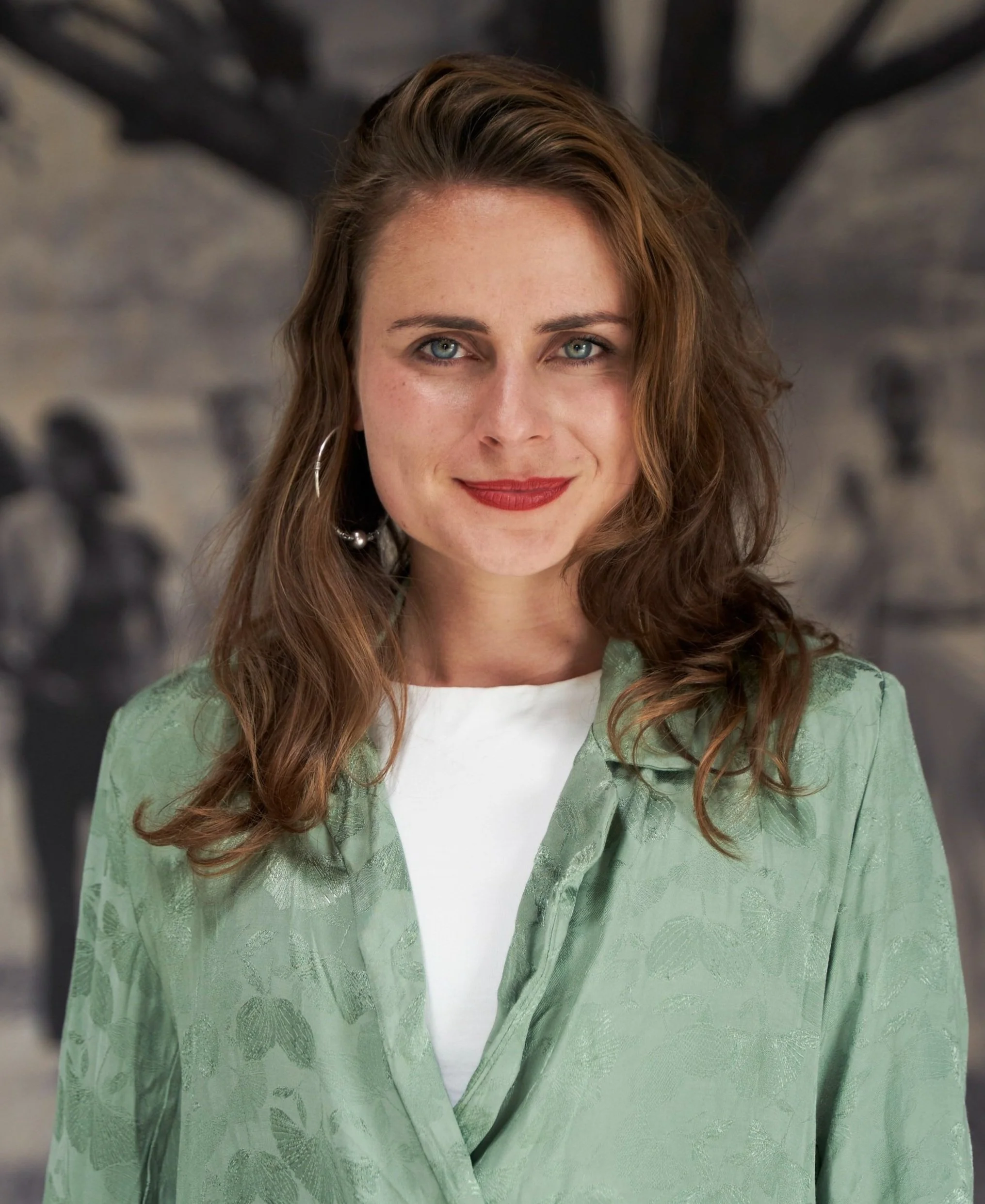 A woman with wavy brown hair, blue eyes, and red lipstick, wearing a light green patterned blouse over a white top, looking at the camera with a slight smile, standing in front of a blurred historical black and white photograph background.