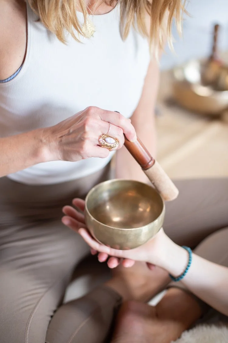 Person playing singing bowl with a mallet while another person holds the bowl. The scene appears to be a peaceful, spiritual or wellness setting.