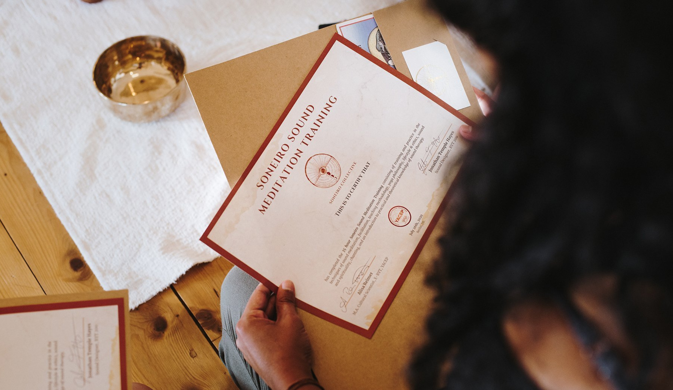 Person holding a certificate for sound therapy meditation training, sitting at a wooden table with a white cloth and a gold cup nearby.
