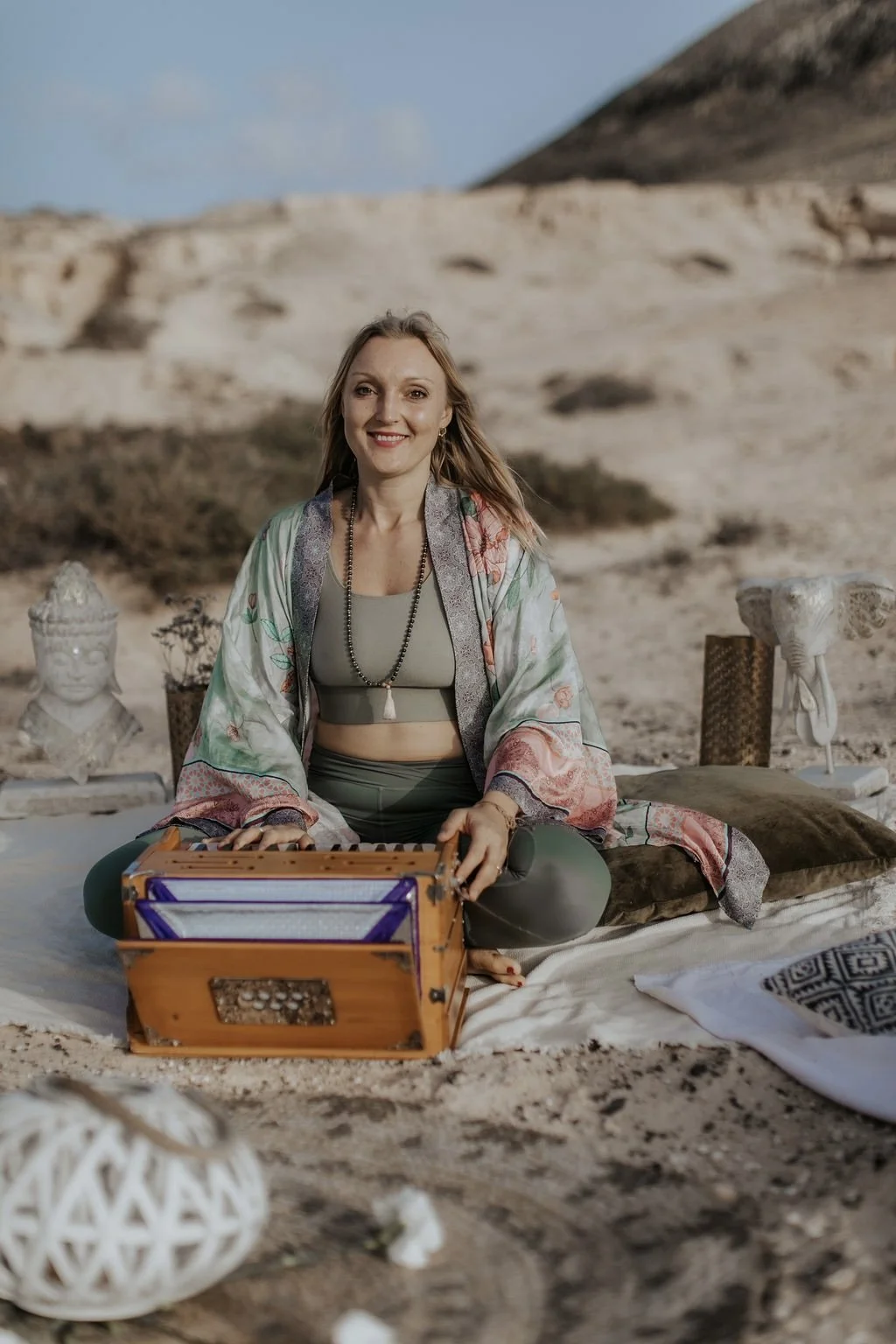 A woman sitting cross-legged on a blanket in a desert landscape, smiling at the camera with sand dunes in the background. She is dressed in a beige crop top, olive green pants, and a colorful shawl, and is surrounded by decorative objects including a wooden box, a bust sculpture, and woven containers.