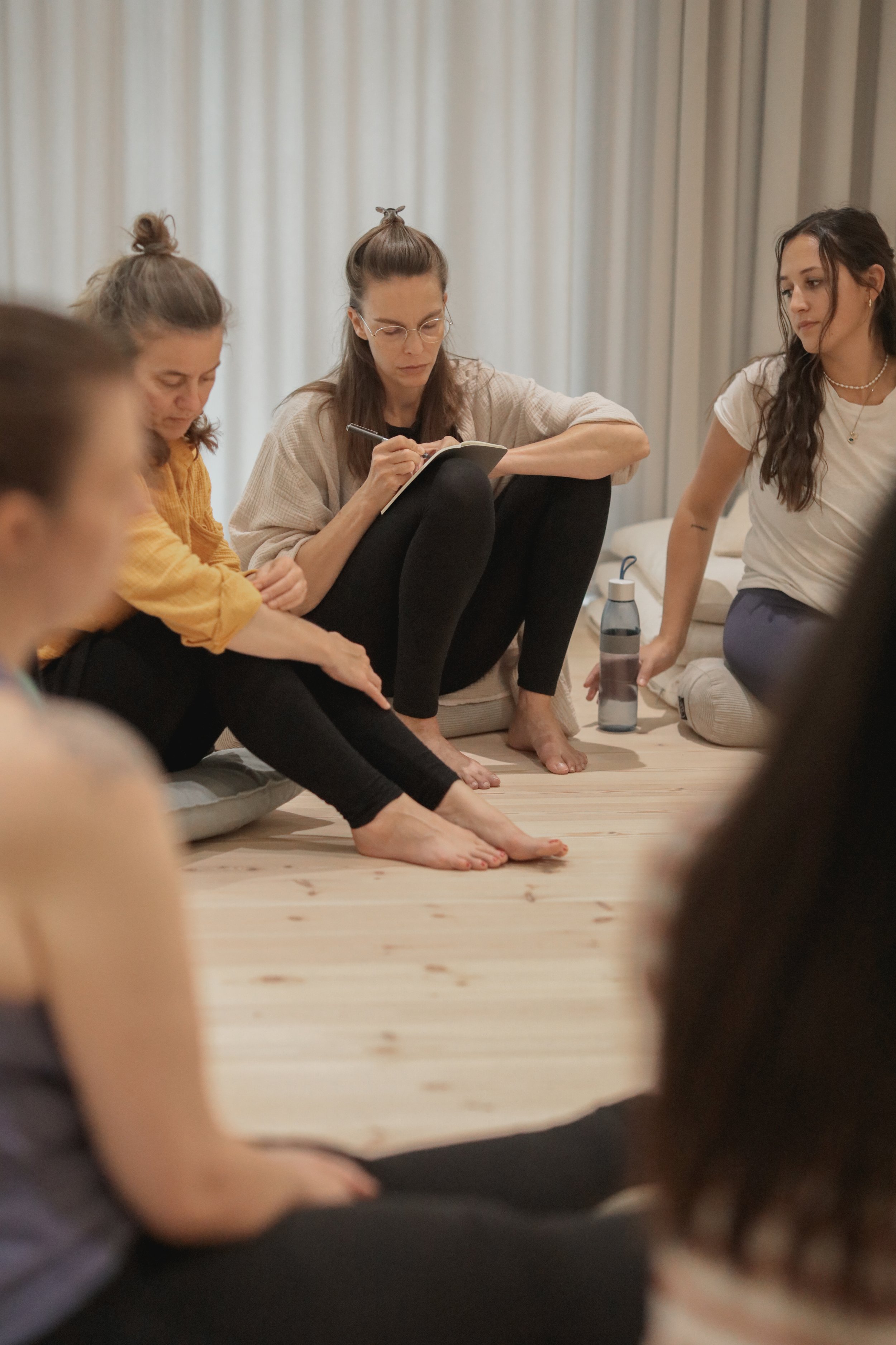 Group of women sitting on the floor in a circle, participating in a discussion or workshop, with one woman taking notes.