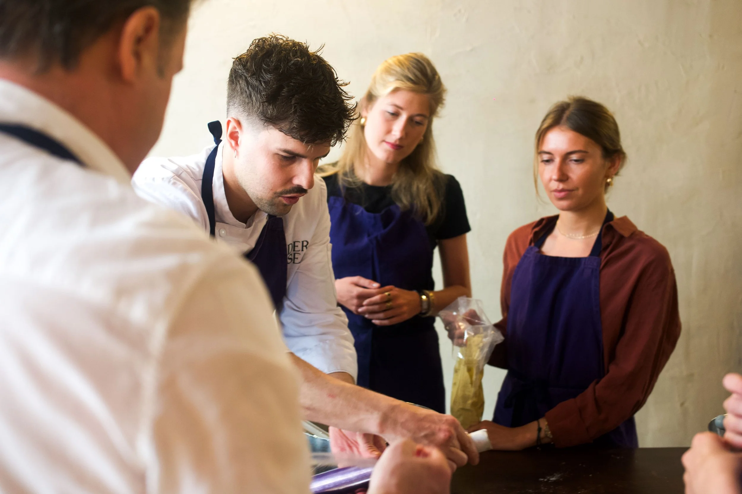 Groupe de personnes en formation culinaire, avec un chef montrant une technique à un groupe de jeunes femmes.