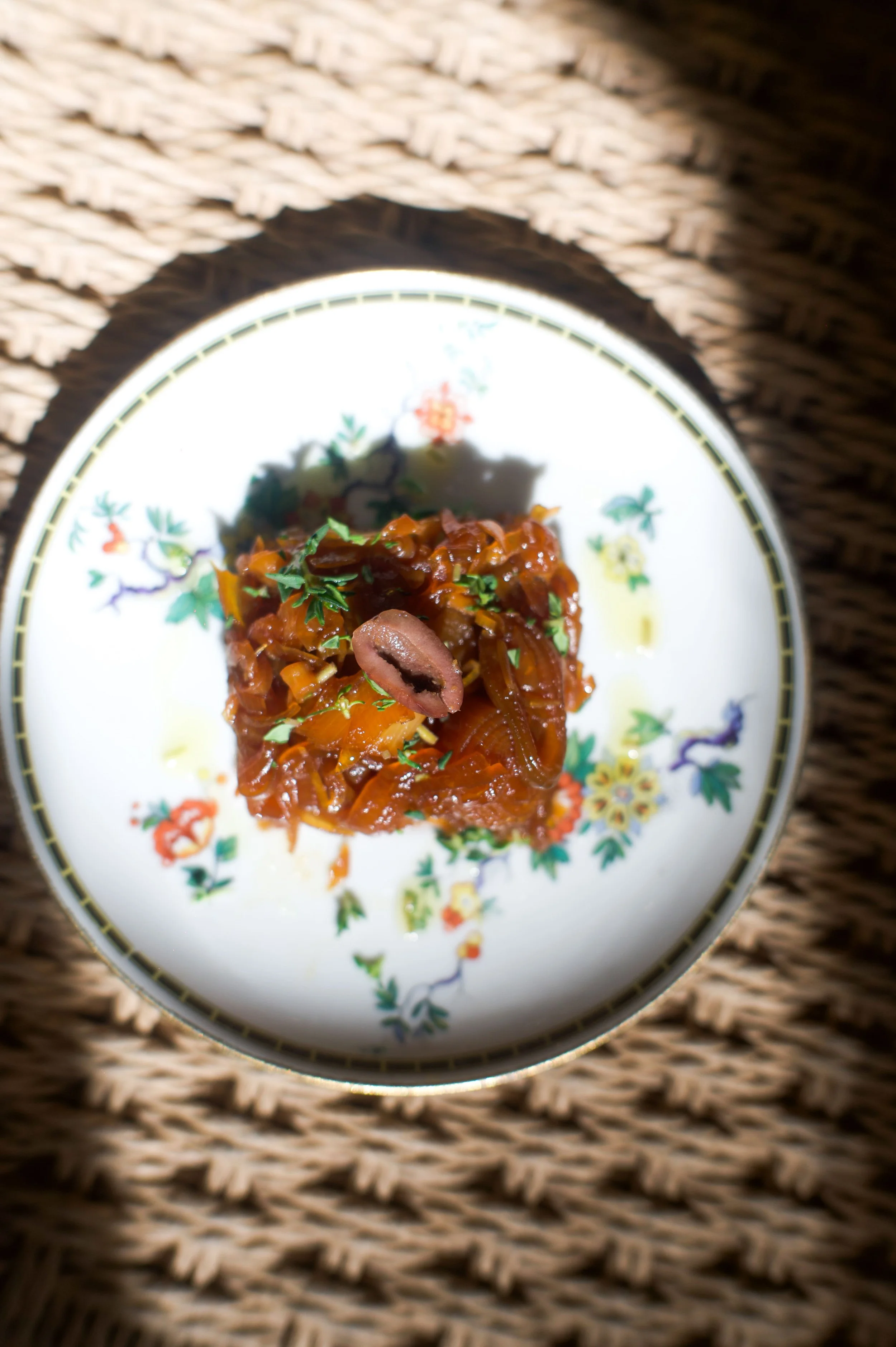 Un plat de cassoulet dans une petite assiette en porcelaine blanche avec un motif floral, posé sur une surface en rotin.