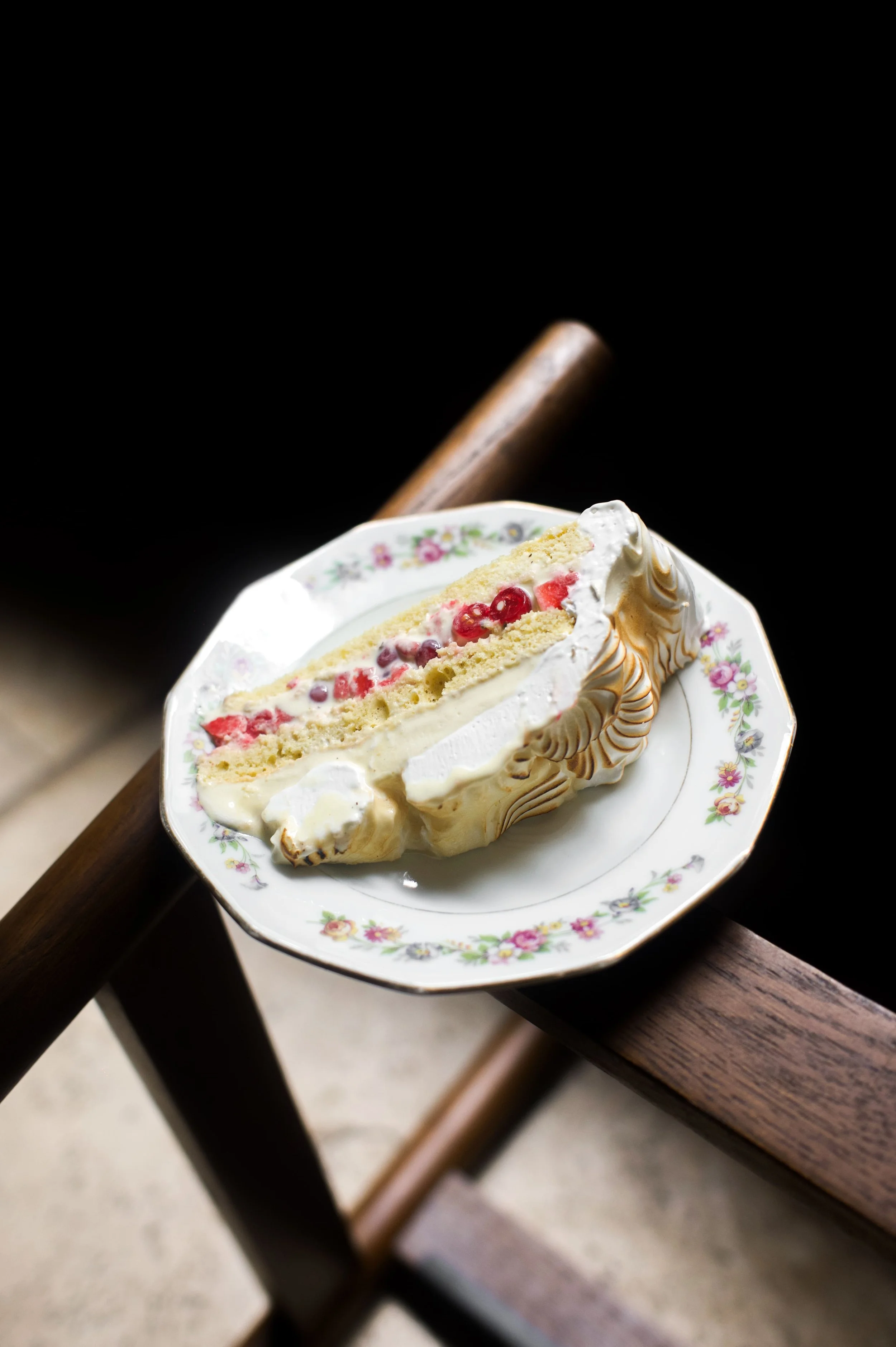 Une part de gâteau à la crème, fruits rouges et génoise sur une assiette en porcelaine blanche avec des fleurs, posée sur une table en bois.