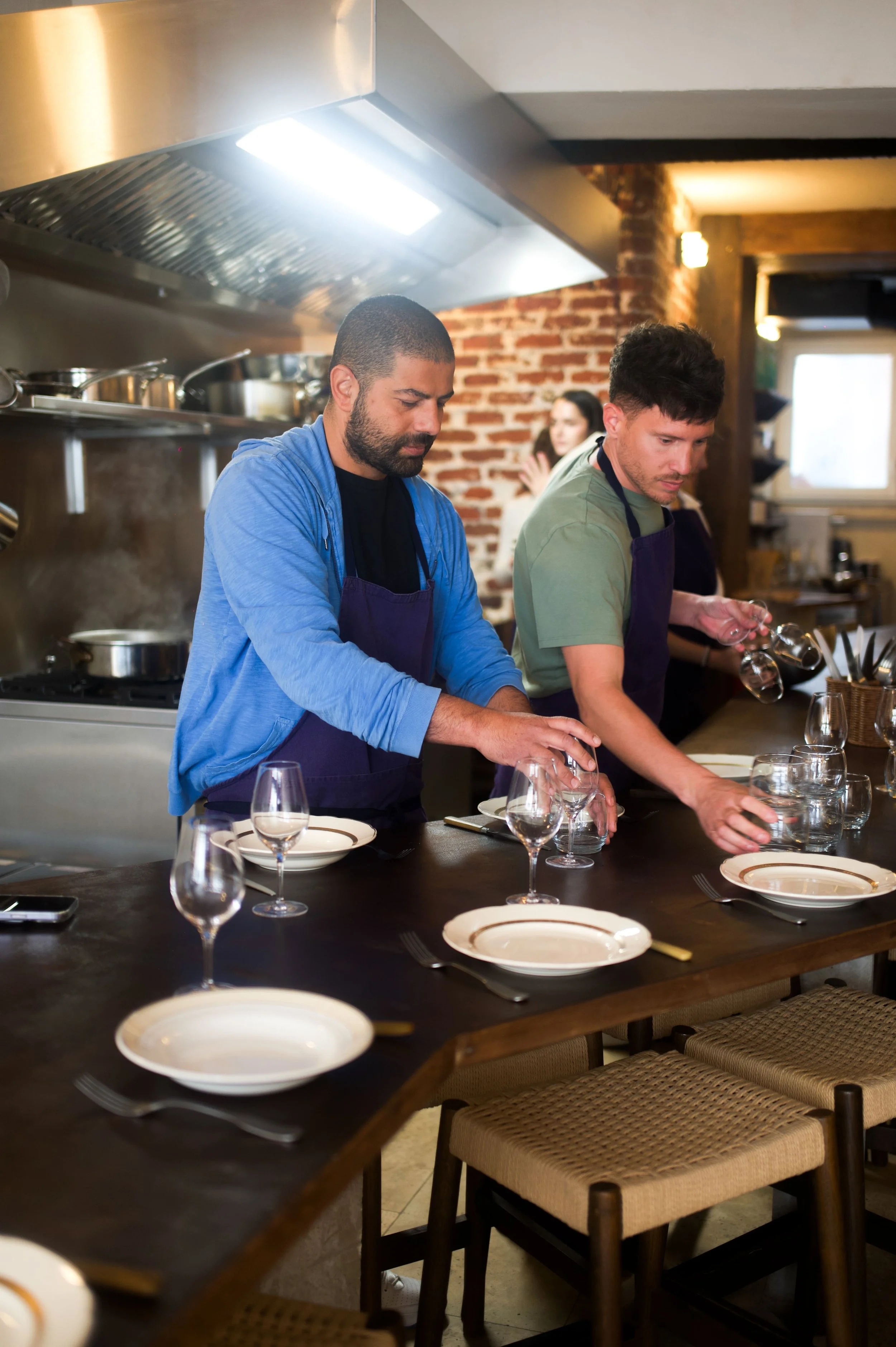 Deux hommes en tabliers préparant une table pour un repas dans une cuisine de restaurant, avec des verres et des assiettes disposés sur la table.