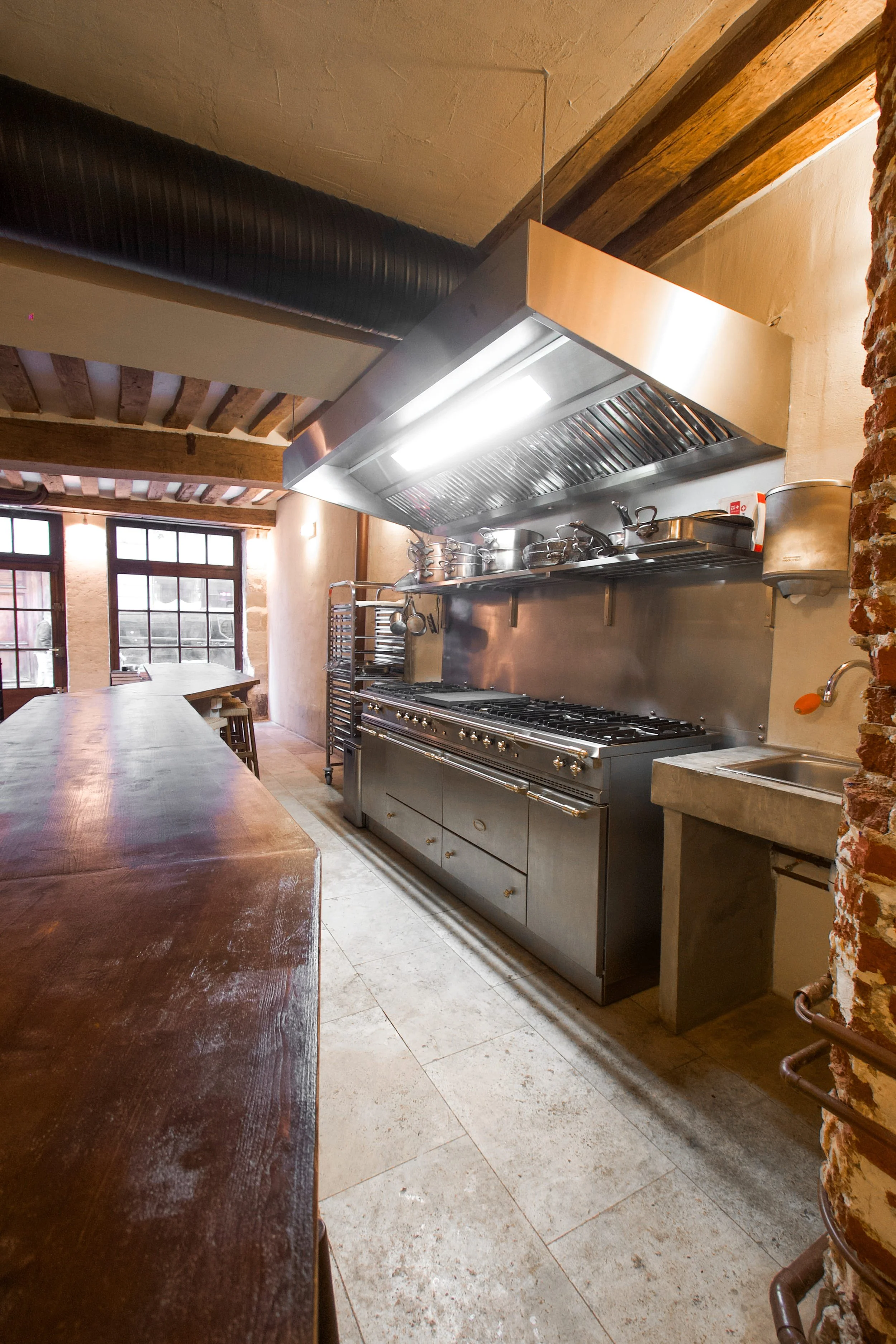 Professional kitchen with large stainless steel gas oven, metal ventilation hood, concrete hand washer and shelf with metal pots and kitchen tools.