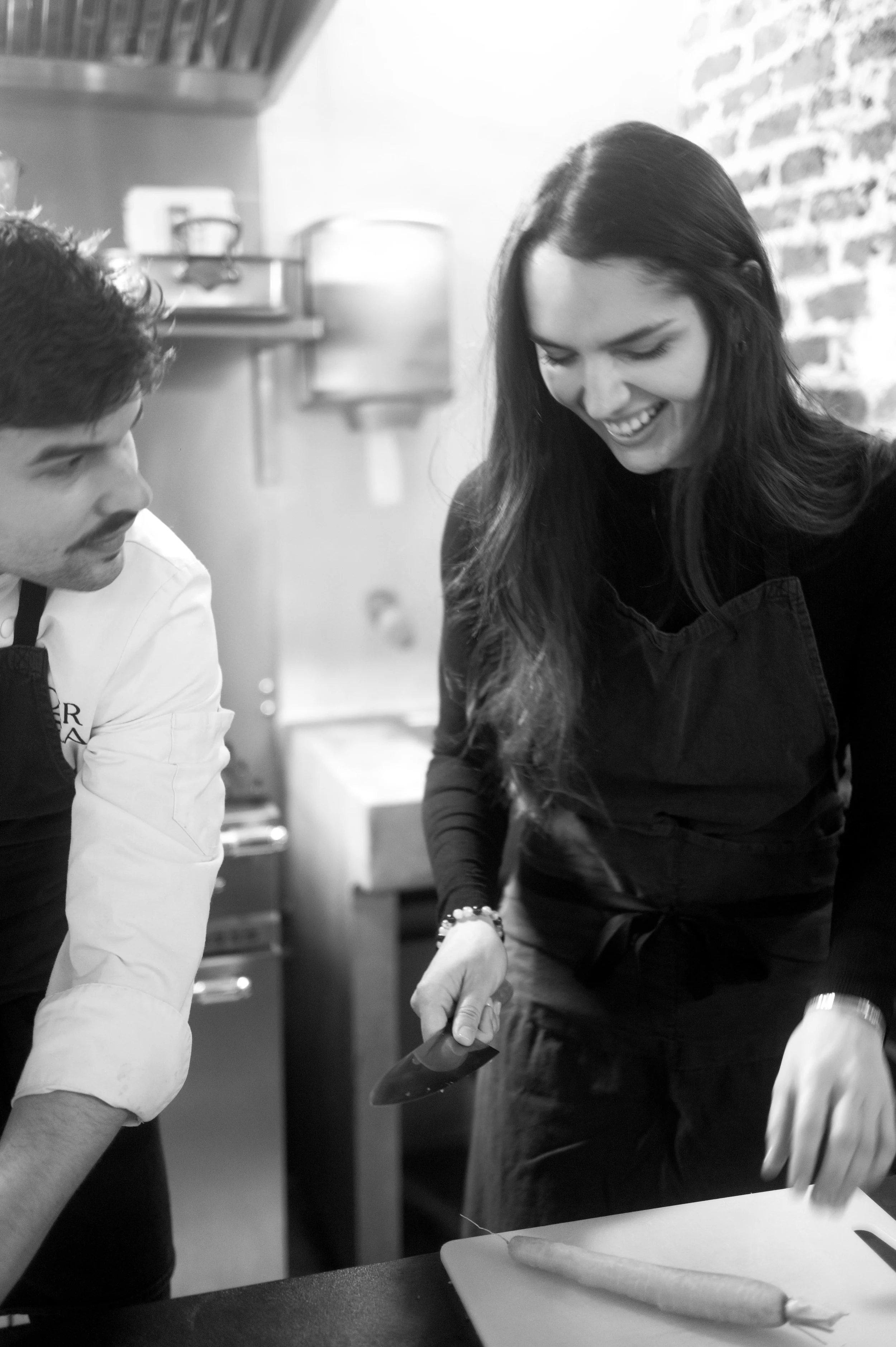 Une femme et un homme en tenue de cuisine, souriants, en train de préparer des légumes dans une cuisine.