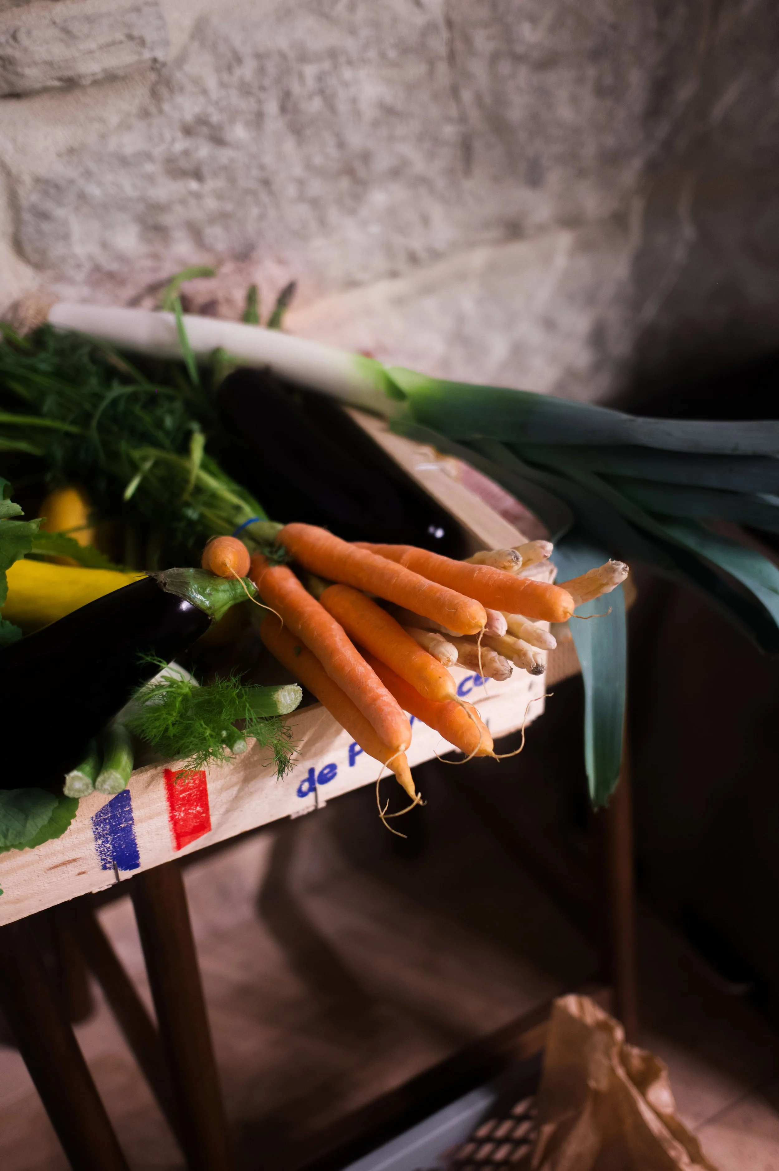 Carottes, aubergines, et poireaux exposés sur une caisse à légumes en bois dans un marché ou une boutique de produits frais.