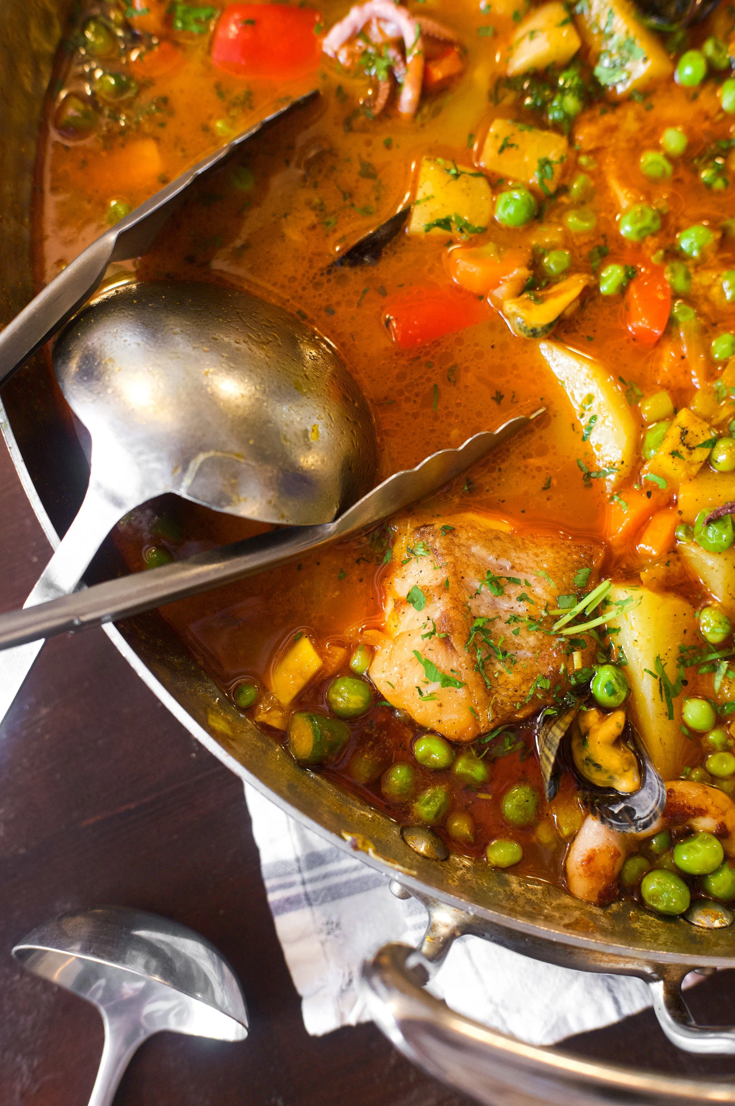 Un plat de soupe ou ragoût avec divers légumes comme pois verts, carottes, pommes de terre, moules, un morceau de poisson, et de la coriandre, dans une poêle en métal.