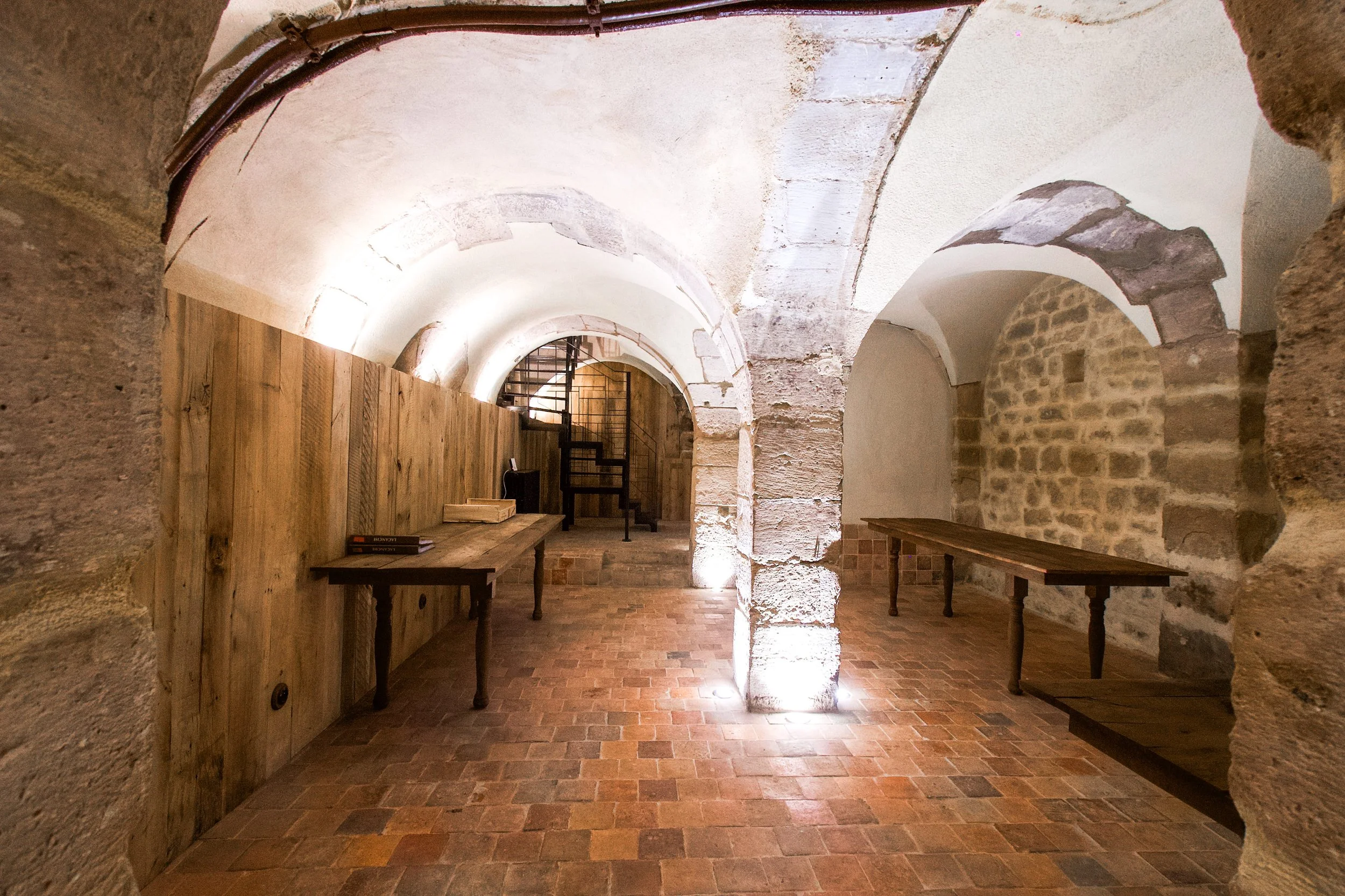 Interior with stone arch, stone and wood walls, brick floor, floor lighting, wooden tables, metal staircase.