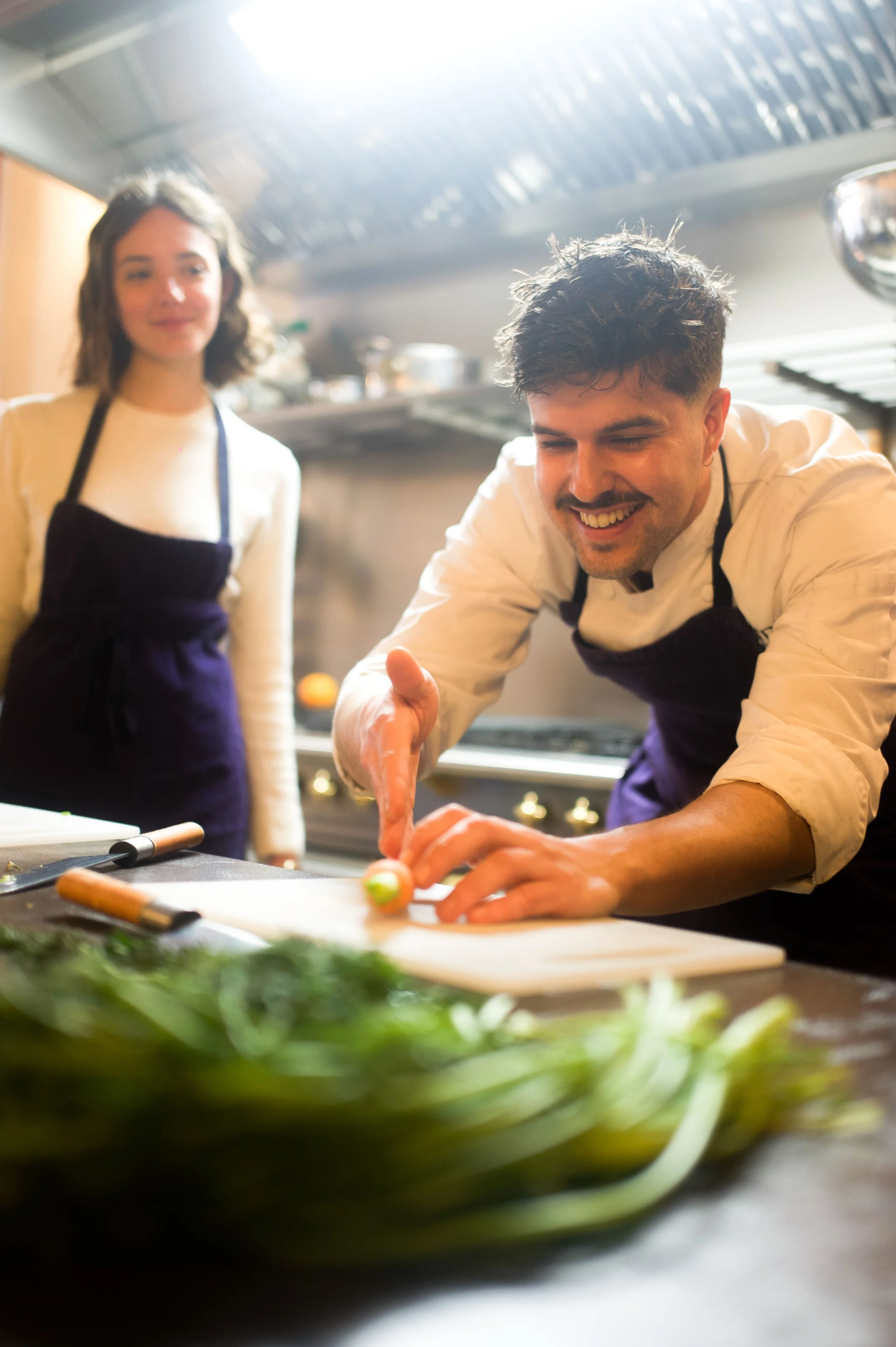 Un chef souriant coupe un légume sur une planche à découper, une assistante observe derrière lui dans une cuisine professionnelle