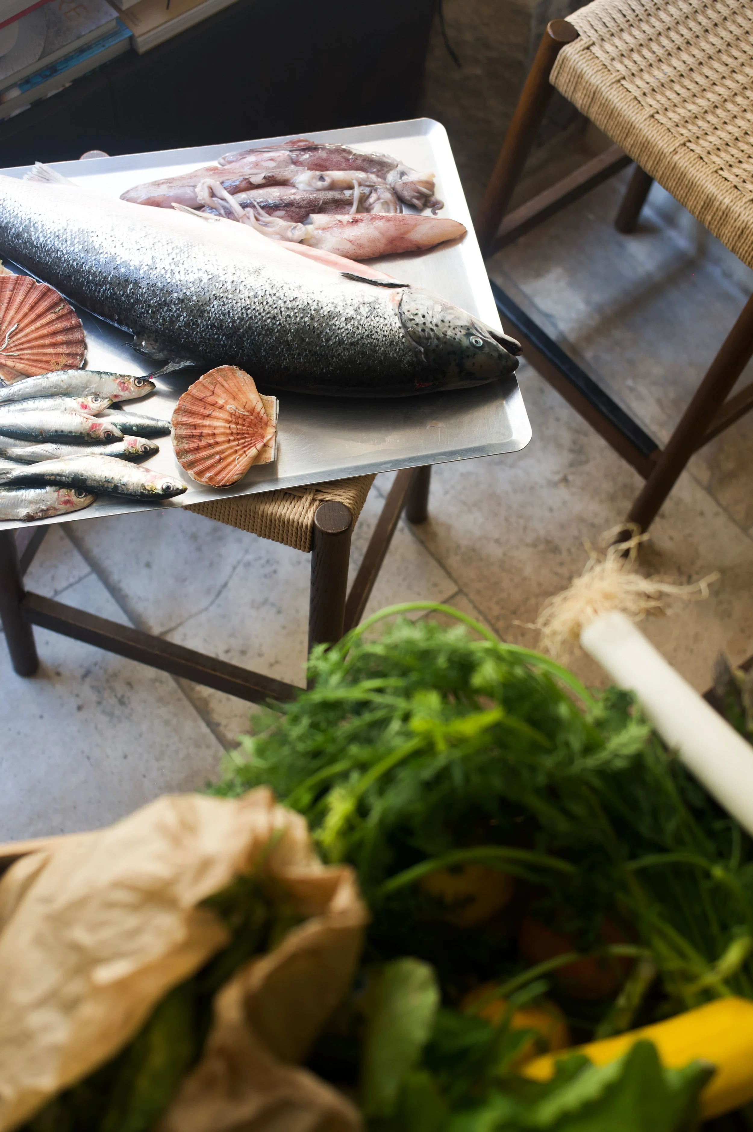 Une planche en métal avec divers poissons, y compris un poisson entier et des calamars, ainsi que des coquilles Saint-Jacques, sur une chaise en bois. En ba­s, il y a des légumes verts.
