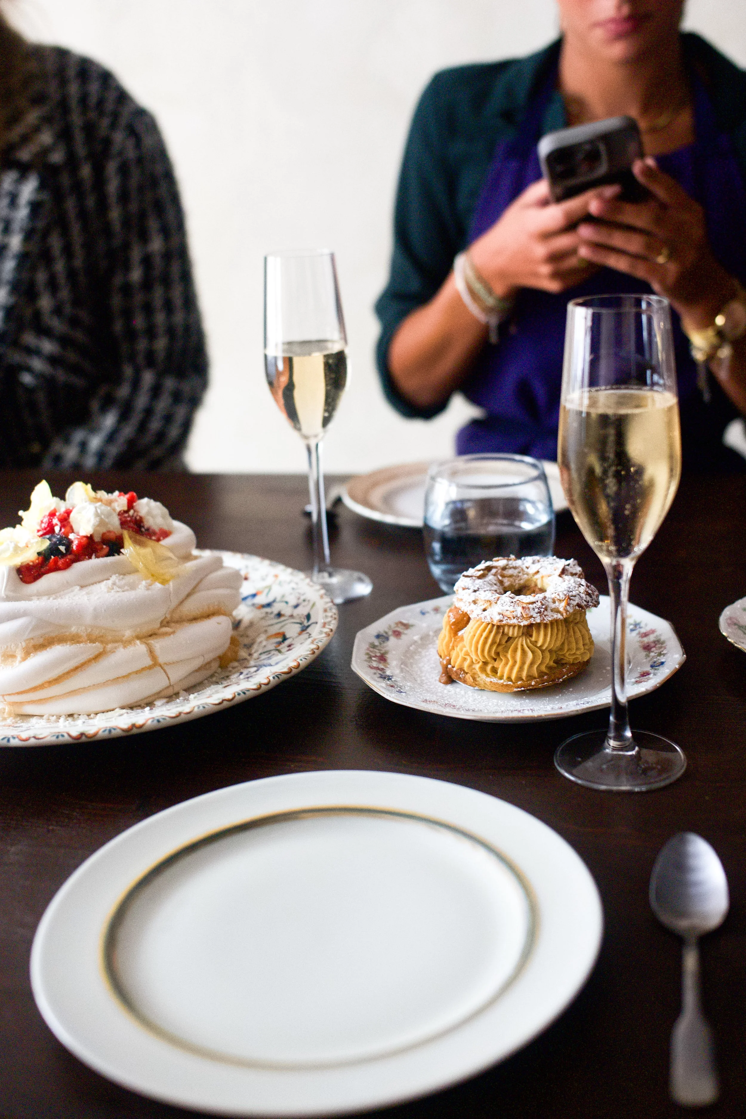 Gâteau pavlova avec des fruits rouges, un choux à la crème, deux flûtes à champagne remplies de champagne, une coupe d'eau, une assiette vide, et des personnes assises autour de la table lors d'une célébration ou fête.
