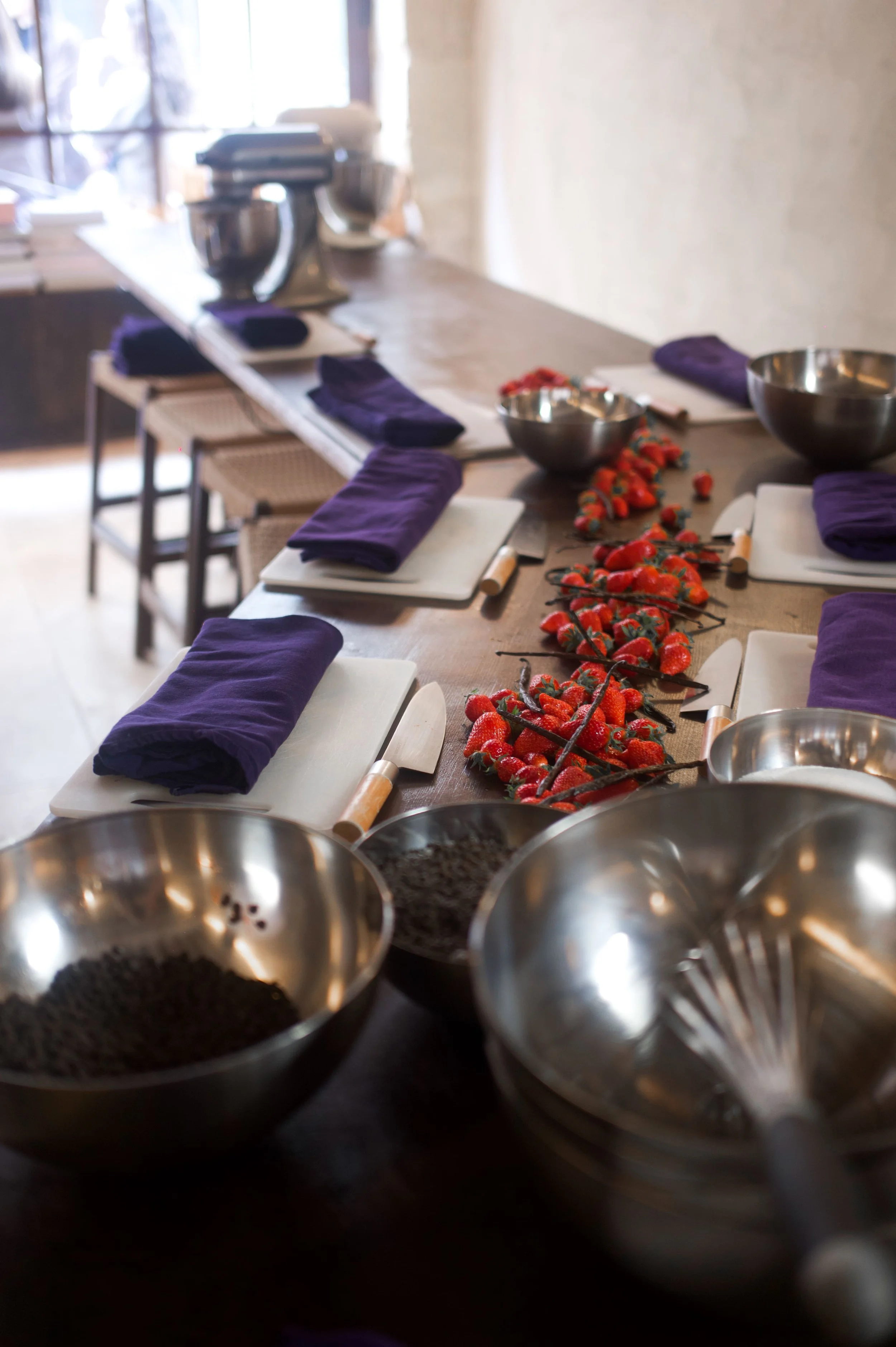 Table de cuisine avec des ustensiles, des serviettes violettes, des bols en acier, une rangée de fraises et des rouleaux à pâtisserie, dans une ambiance lumineuse.