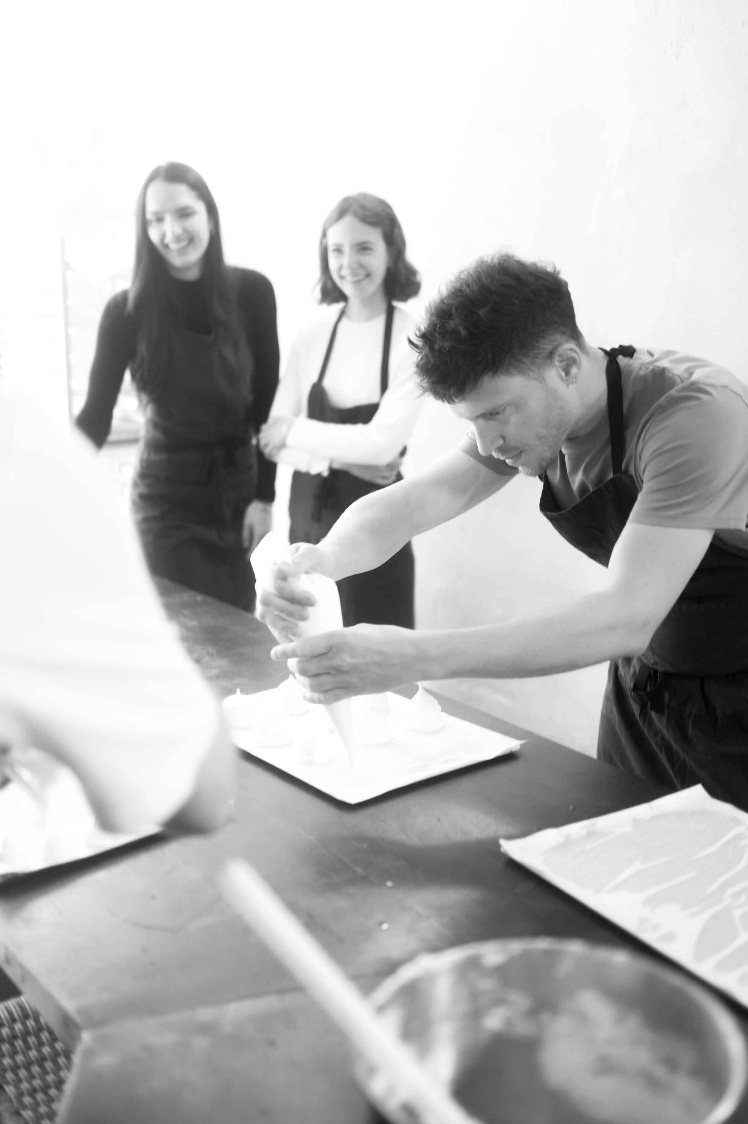 Groupe de trois personnes dans une cuisine ou un atelier de pâtisserie, avec un homme utilisant une poche à douille, et deux femmes observant, toutes portant un tablier.