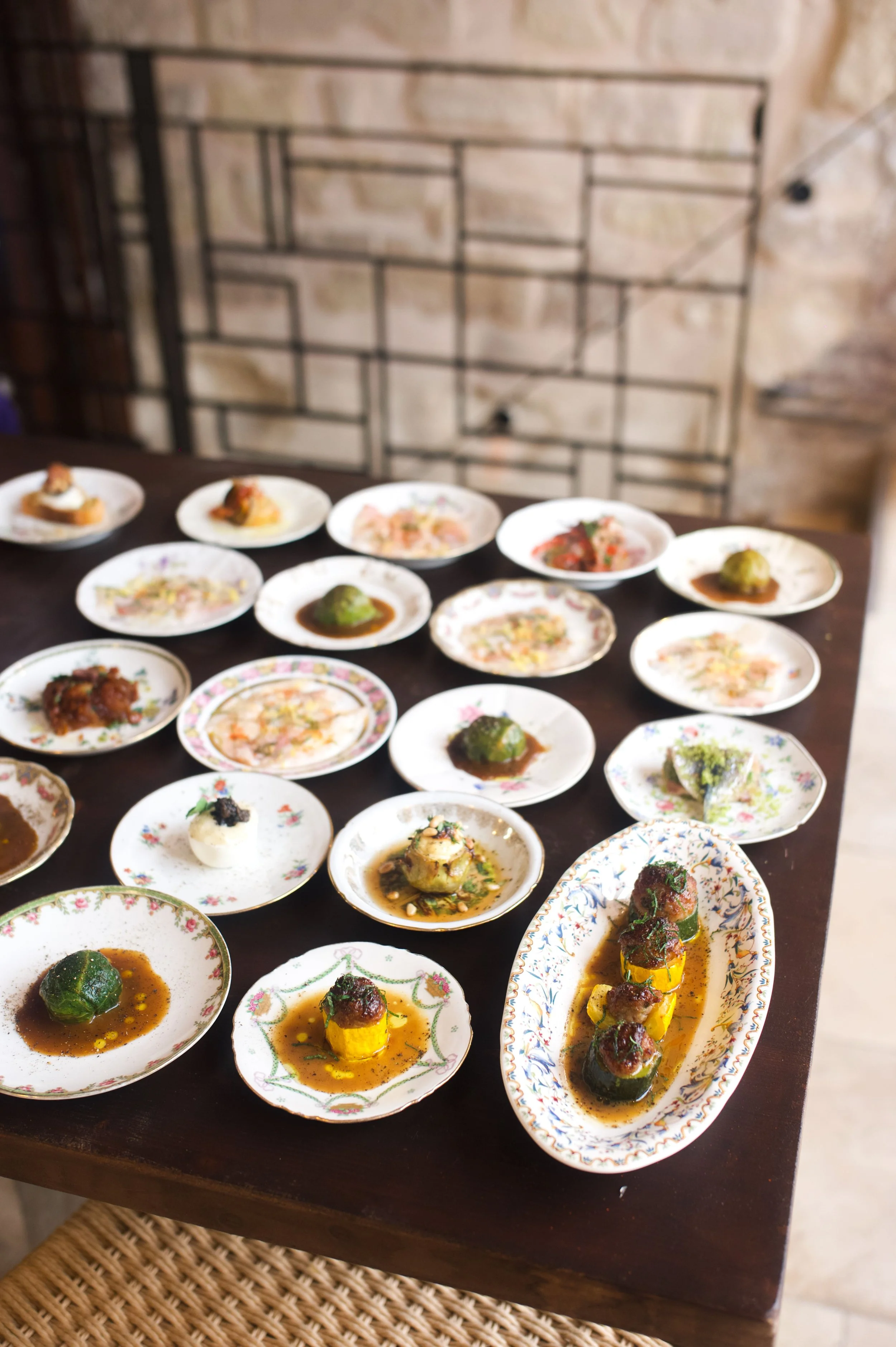 Une variété de petits plats gastronomiques présentés sur plusieurs assiettes en porcelaine. Le fond est une surface en bois, avec un mur en pierre en arrière-plan.