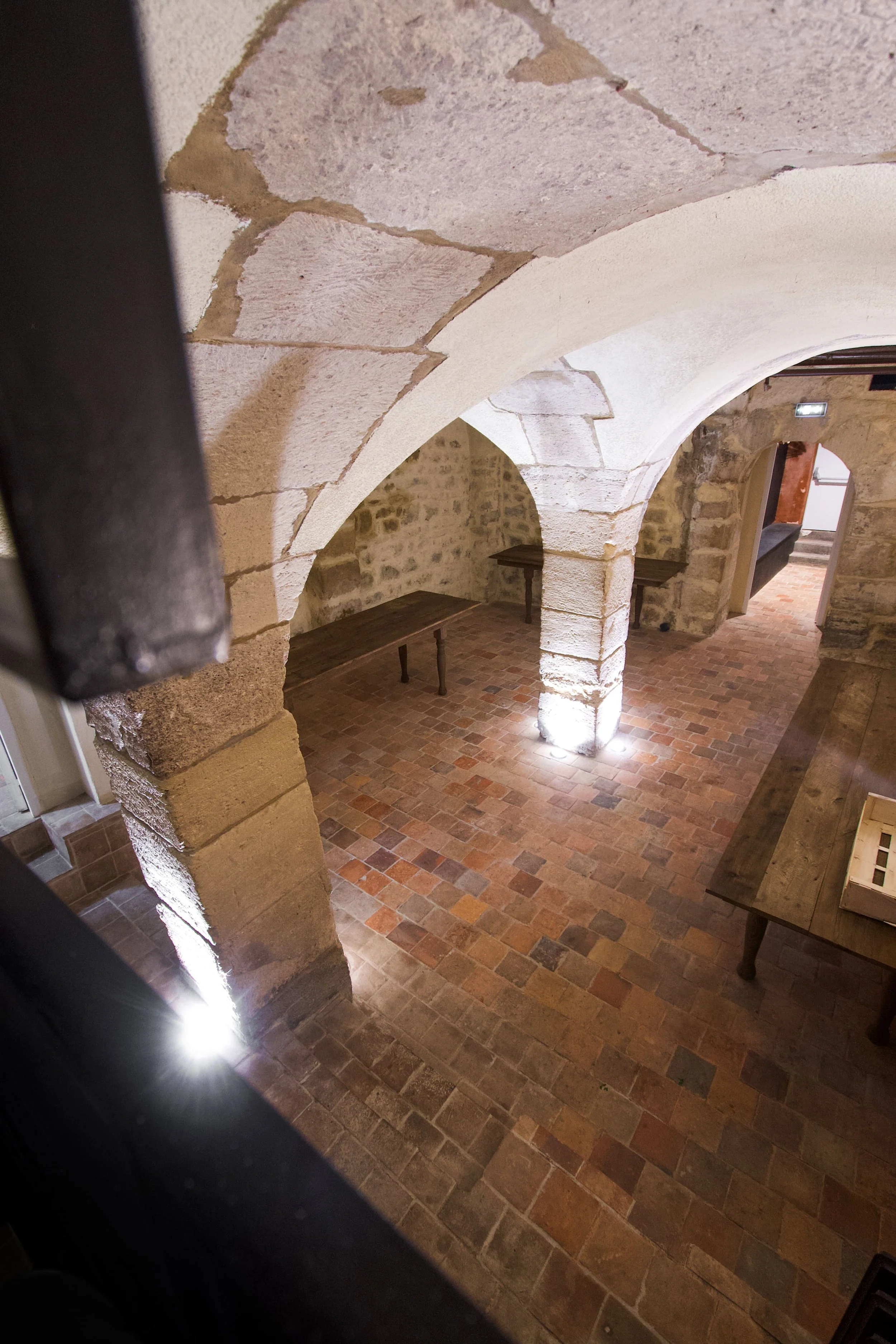 View from above of a room with stone walls, arches and a terracotta tile floor, with wooden tables in a medieval or rustic ambiance.