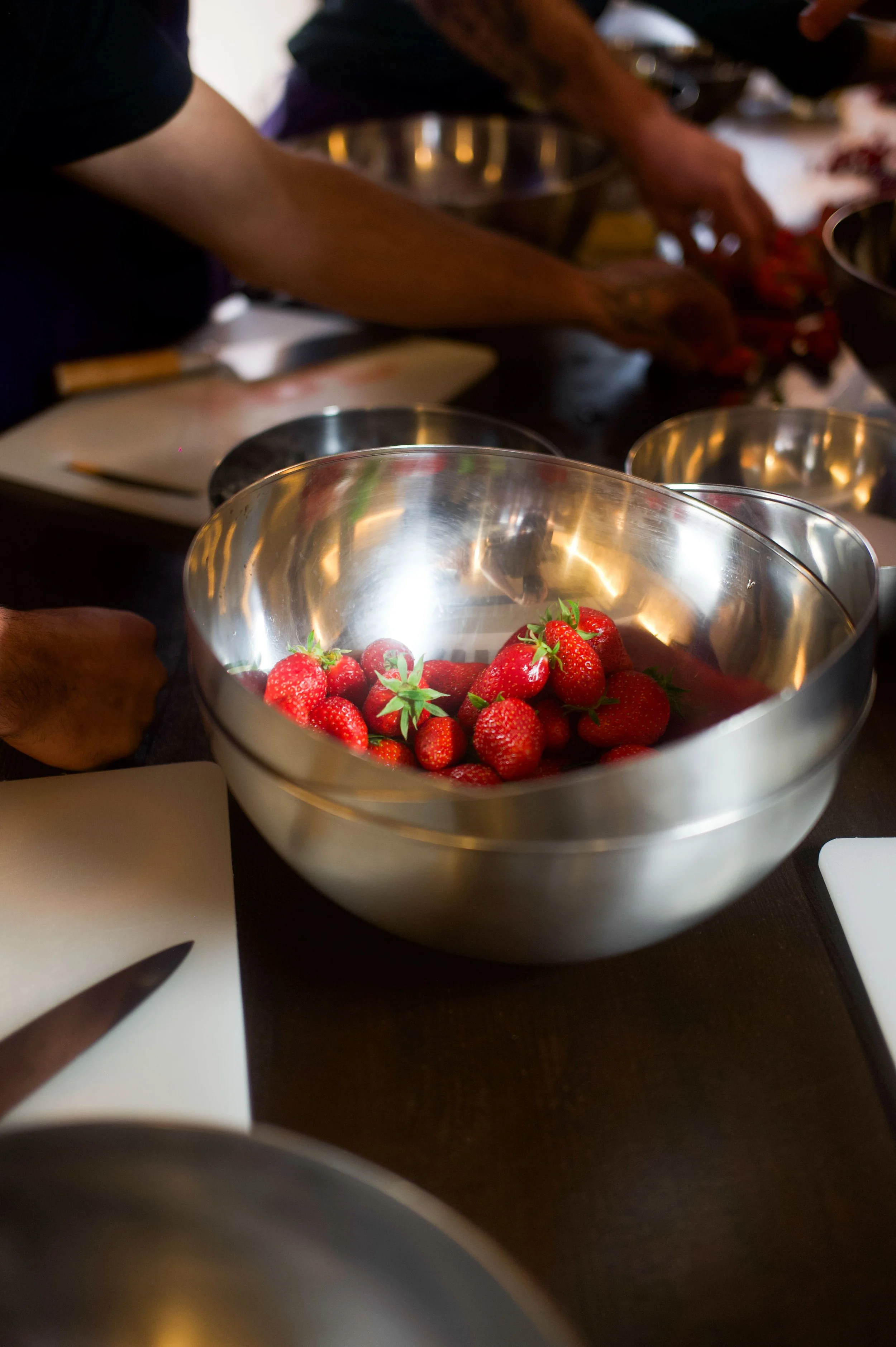 Bol de fraises fraîches sur une table de cuisine avec des personnes préparant des ingrédients en arrière-plan.