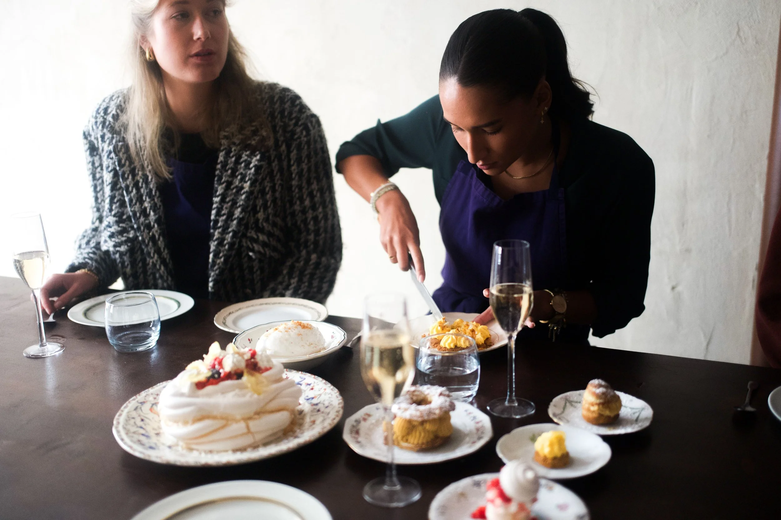Deux femmes autour d'une table avec des desserts et des flûtes de champagne, l'une coupe un gâteau pendant que l'autre regarde.