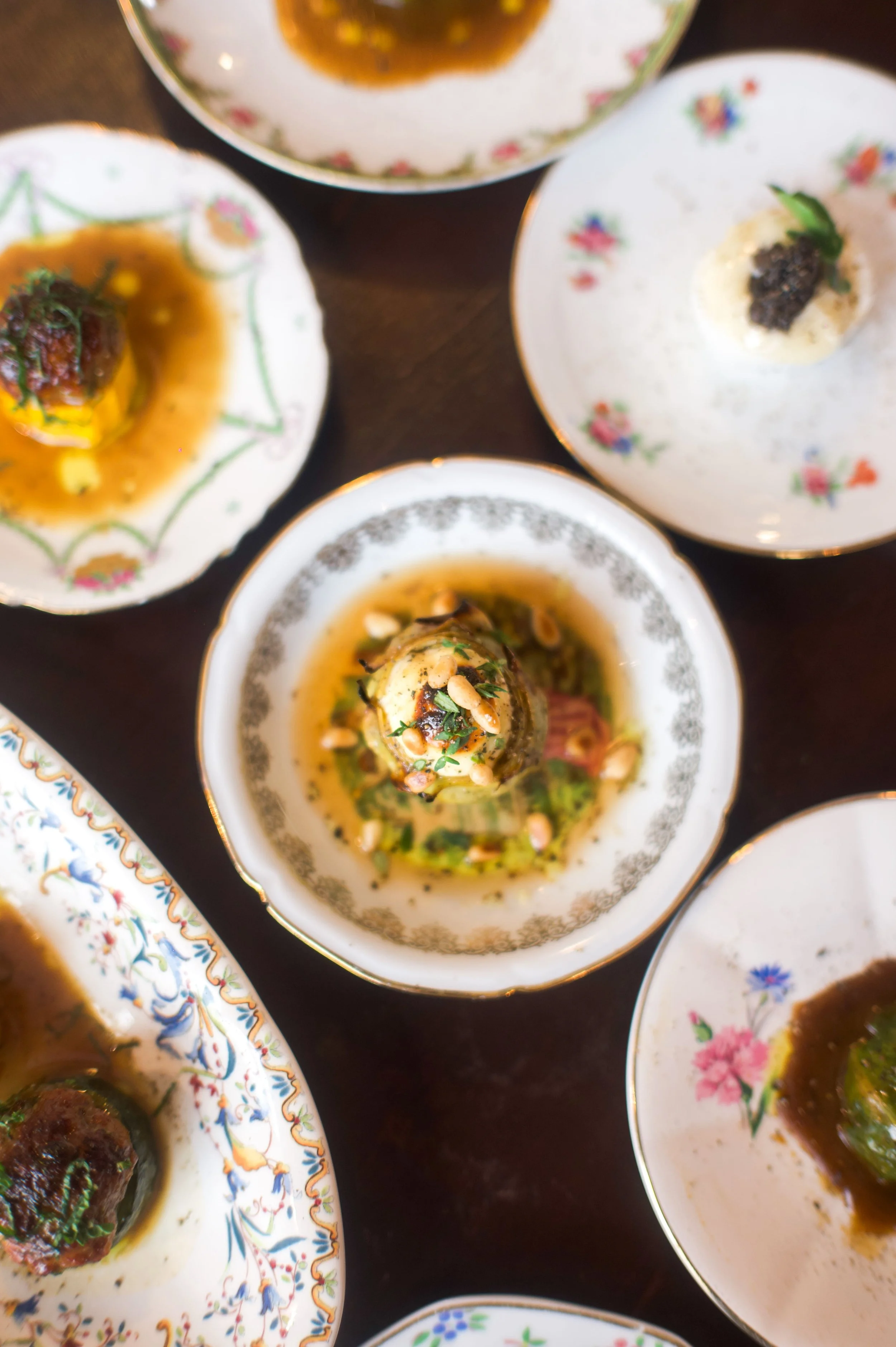 Plats de cuisine élégants dans de la vaisselle délicate avec motifs floraux, comprenant des aliments en sauce, des garnitures de noix, et divers ingrédients sophistiqués, servis sur une table en bois.