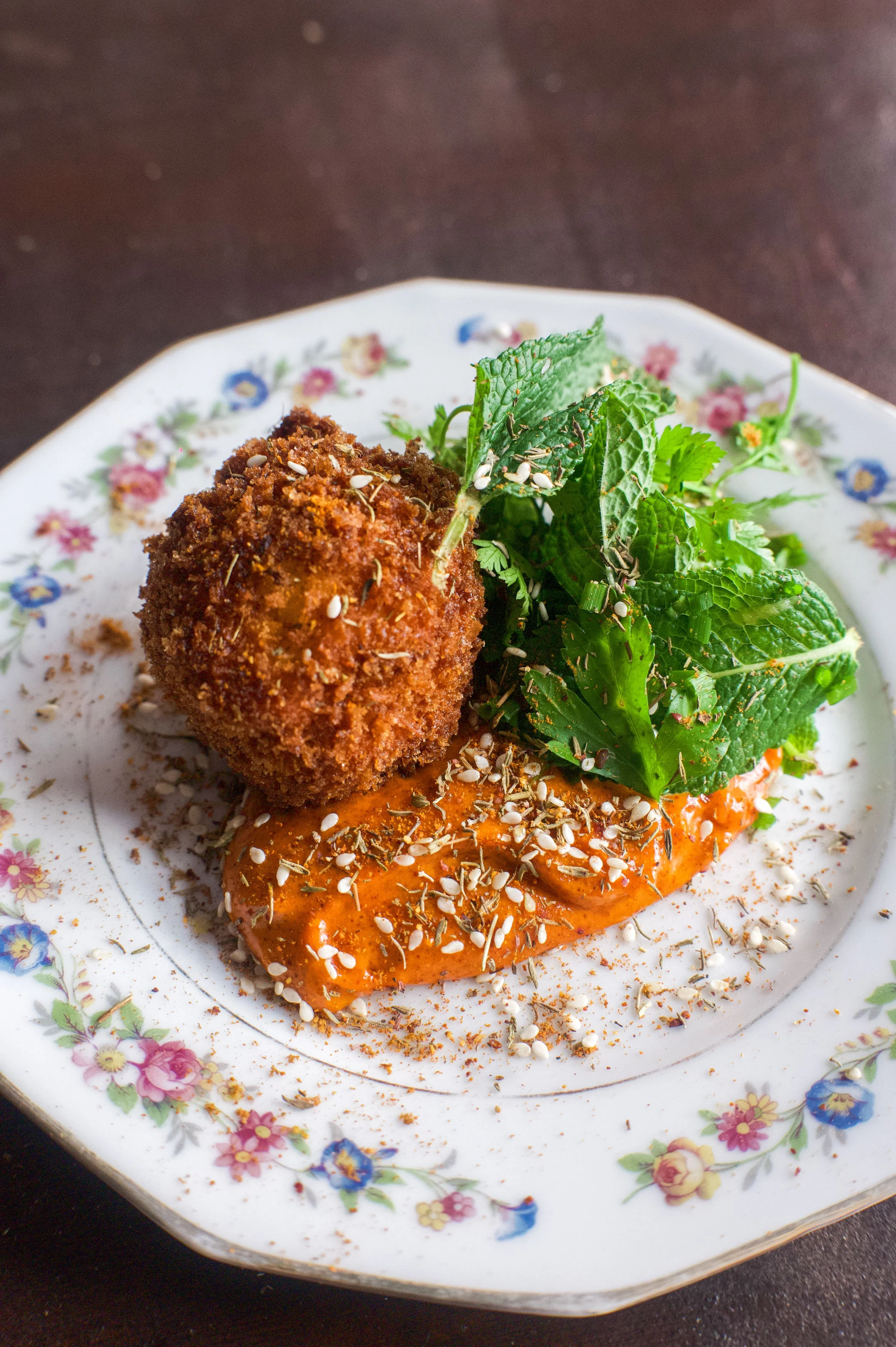 Plat de nourriture avec une boule de croquette, une sauce orangée, des graines de sésame, des herbes et des feuilles de menthe, présenté sur une assiette à fleurs.