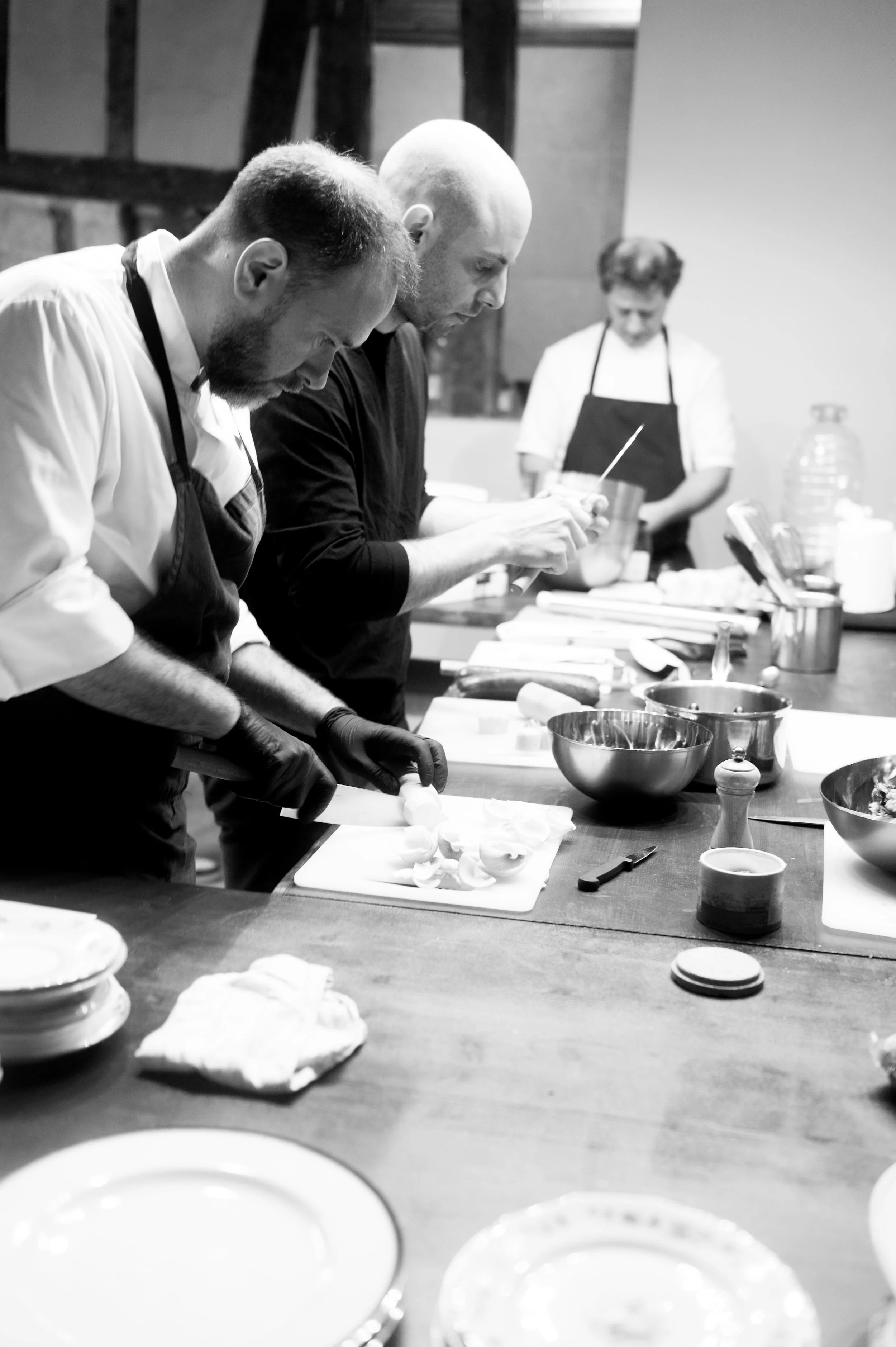 Trois chefs en cuisine préparant des plats dans une cuisine professionnelle, équipés de tabliers et d'outils de cuisine, travaillant sur un plan de cuisson.