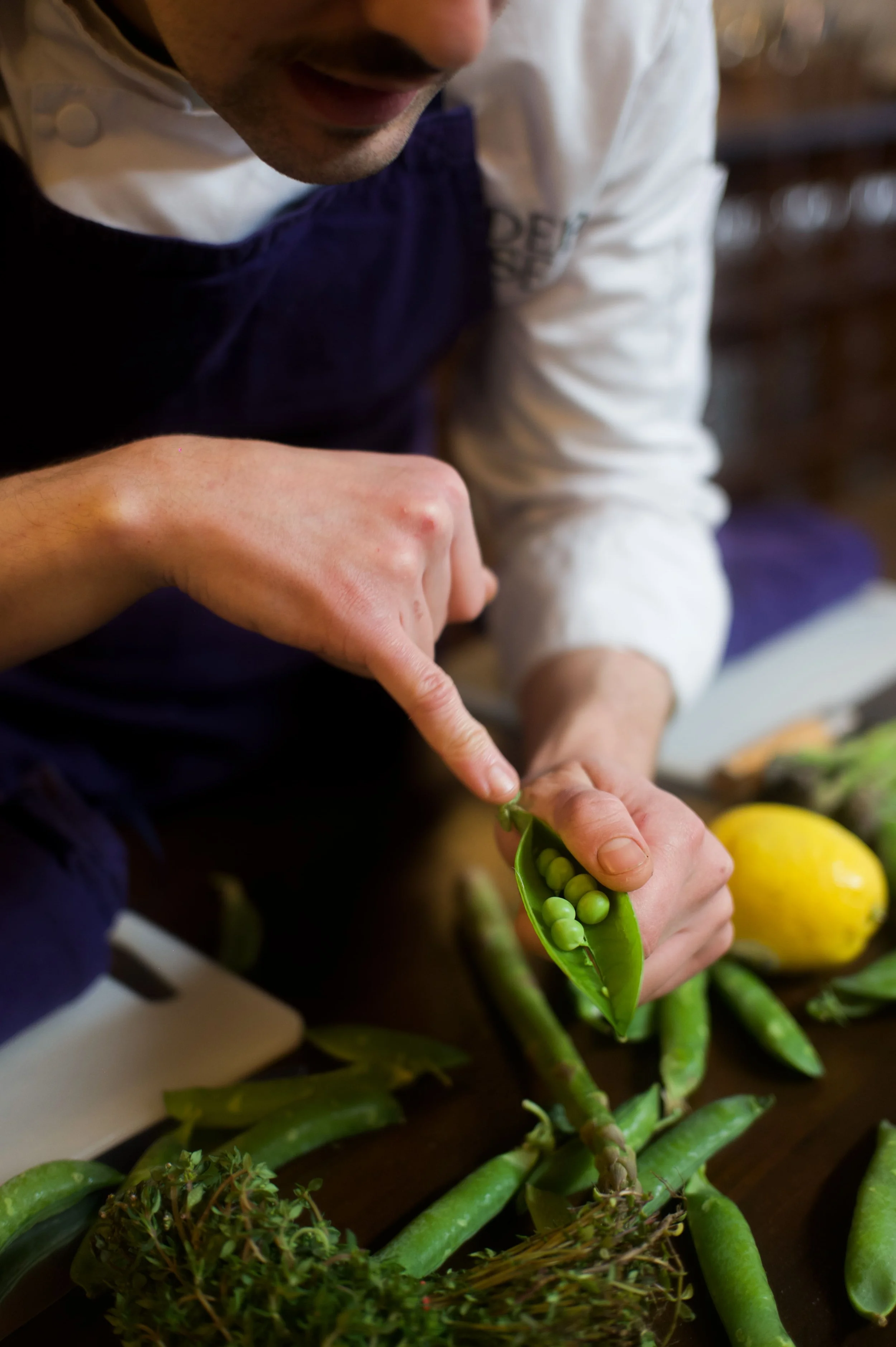 Un chef en train d'ouvrir une gousse de petits pois verts sur une table entourée de légumes, dont un citron jaune et des haricots verts.