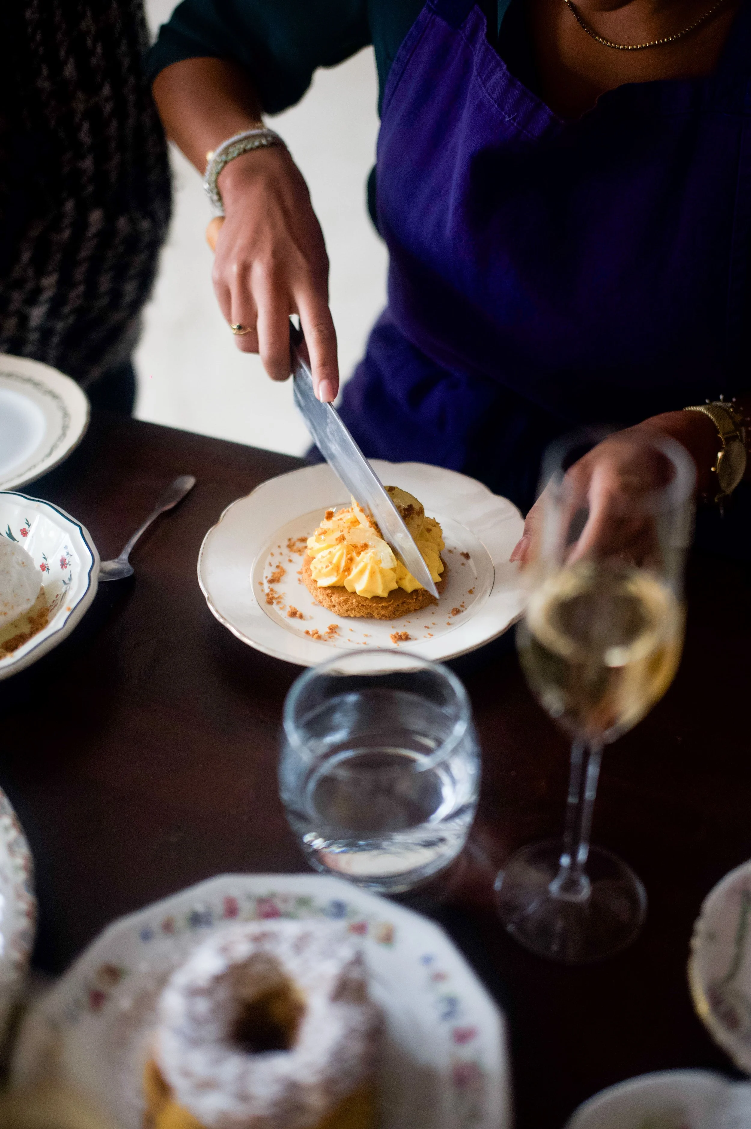 Une personne coupe un dessert à la crème sur une assiette à table, entourée d'autres plats, une verre d'eau et un verre de champagne.