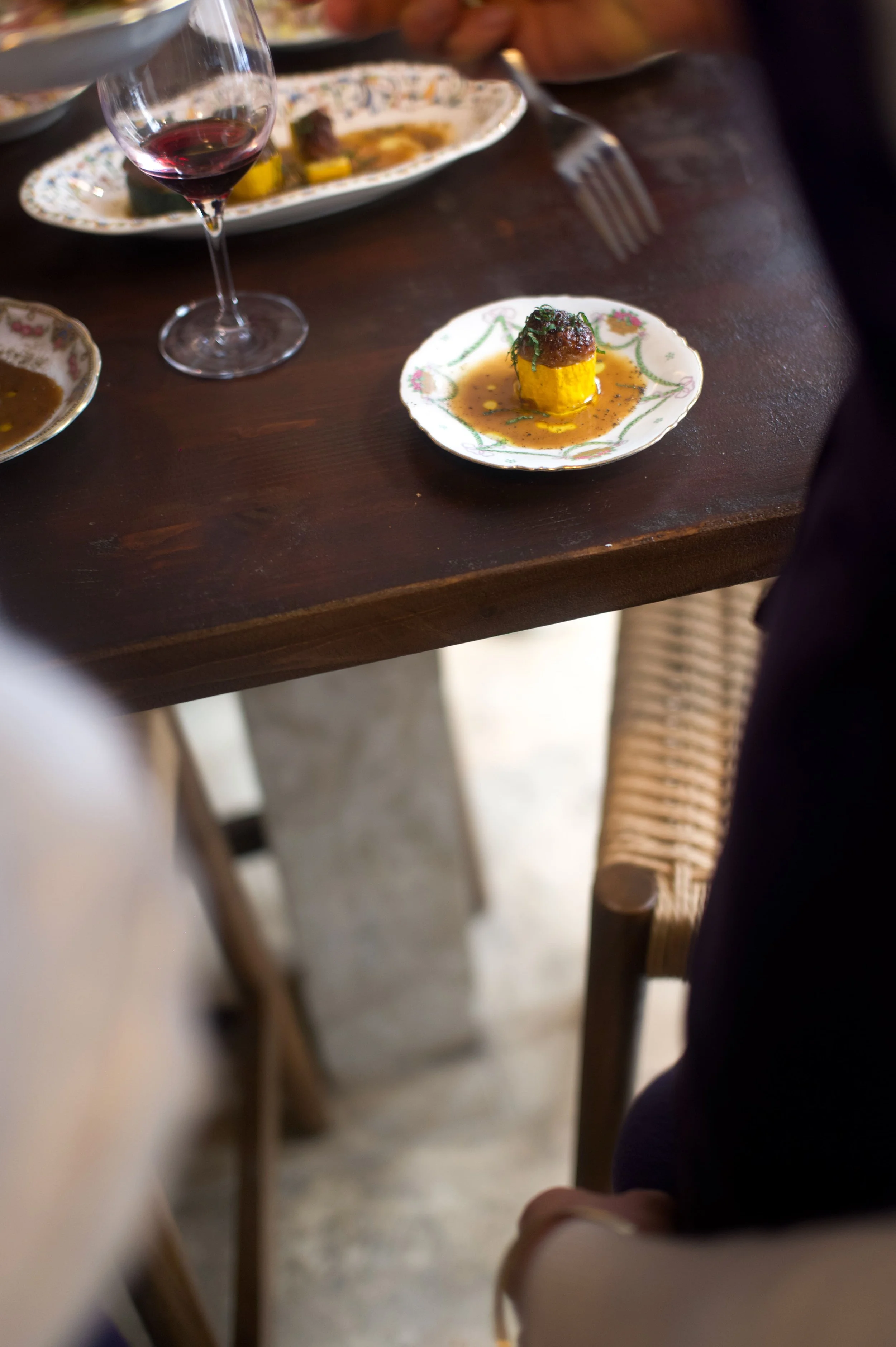 Une assiette avec une portion de nourriture miniature, probablement une amuse-bouche ou un apéritif, comprenant un morceau de viande ou de légumes recouvert d'une sauce, sur une petite assiette décorative, à côté d'un verre de vin rouge. En arrière-p
