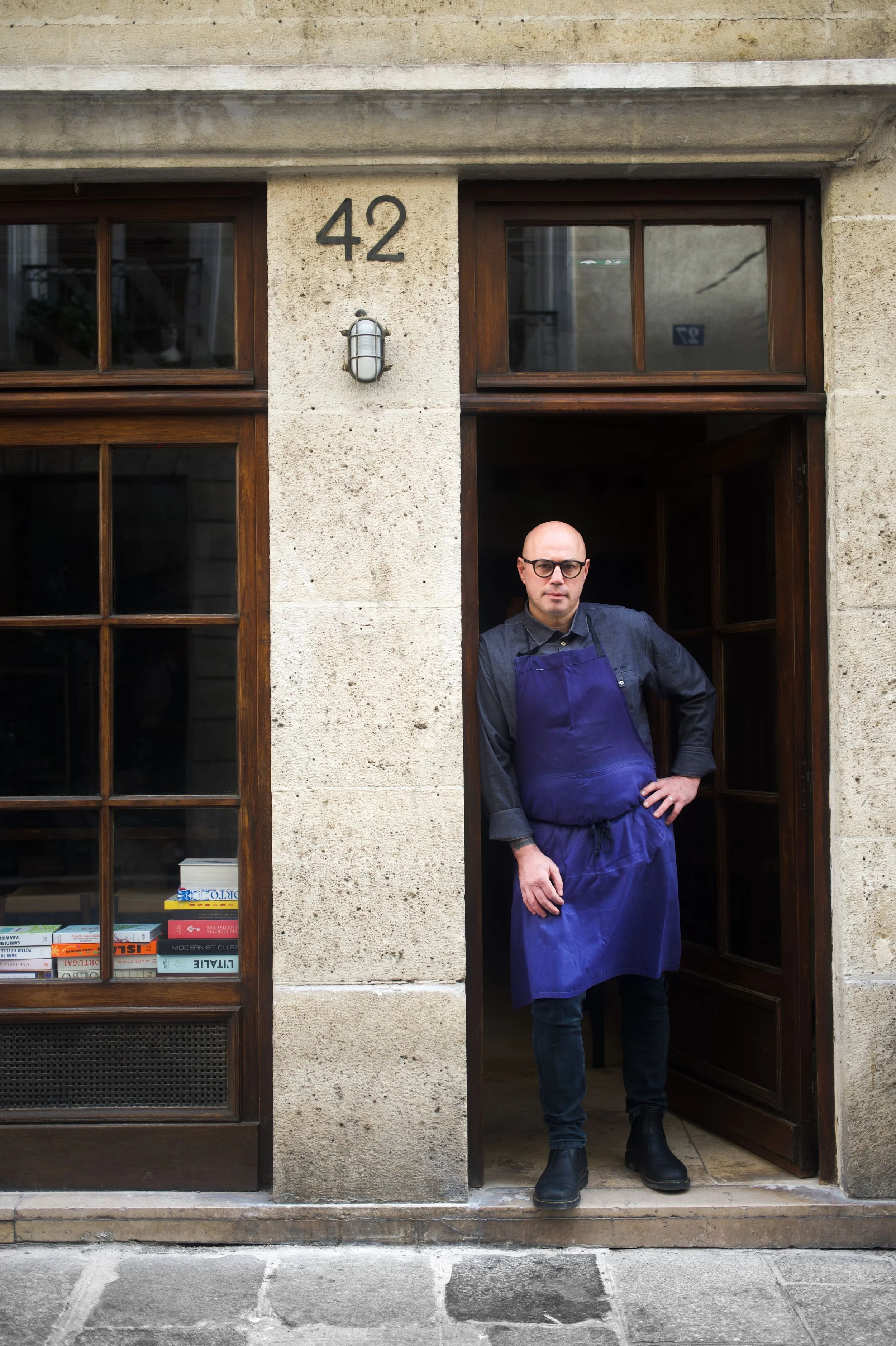 Un homme portant un tablier bleu marine se tenant dans l'embrasure d'une porte en bois, devant un bâtiment en pierre avec le numéro 42 au-dessus. Il porte des lunettes et pose avec une main sur la hanche.