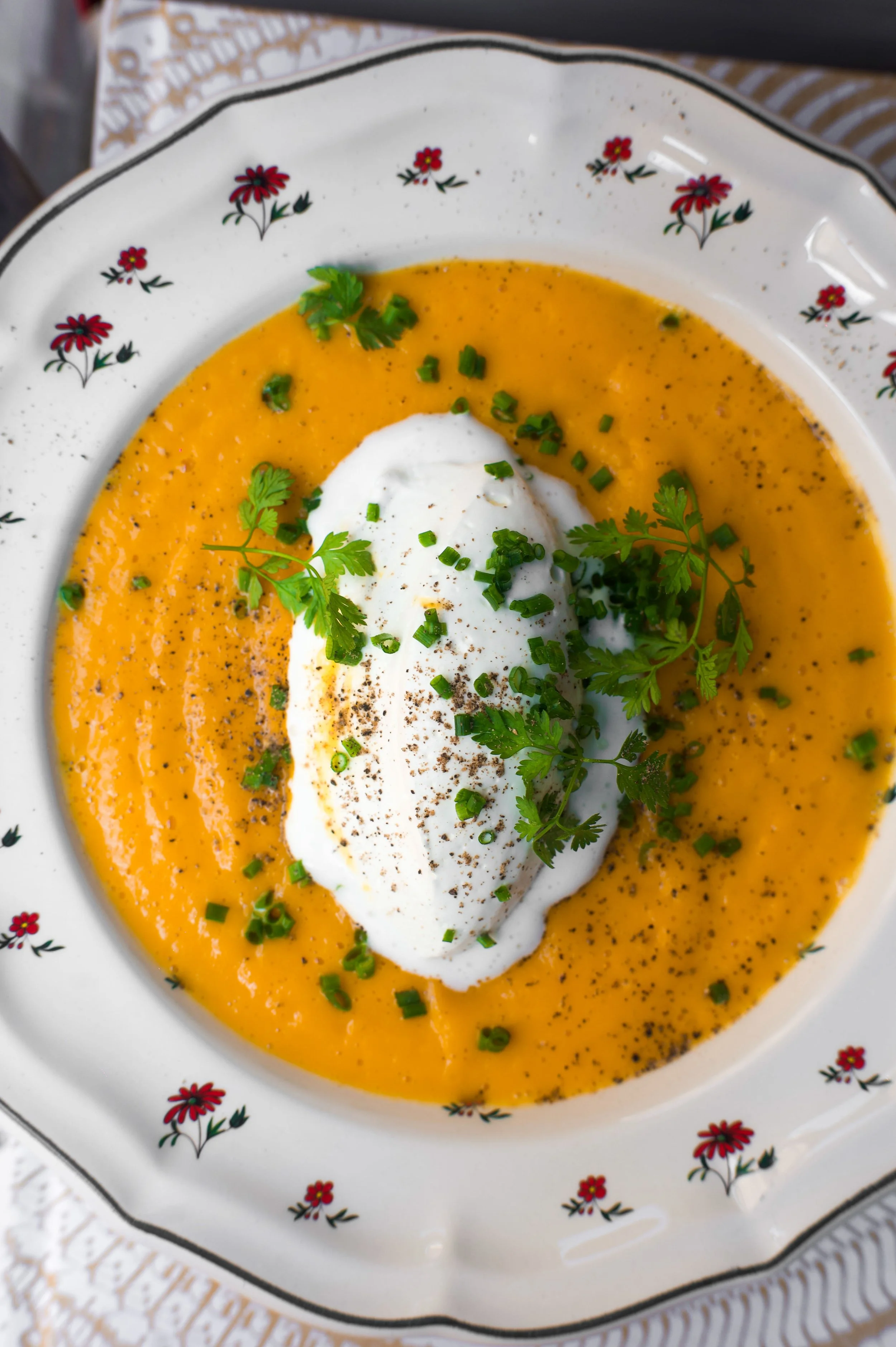Soupe de carottes avec une cuillère de crème, garnie de persil, servie dans une assiette blanche avec des fleurs rouges.