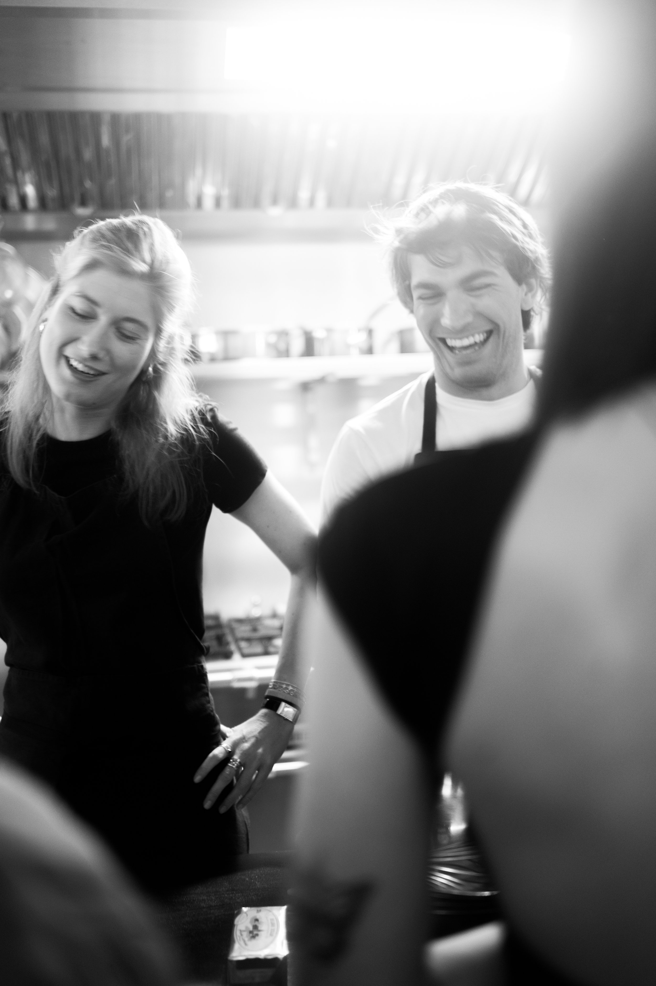 Deux jeunes adultes souriants, un homme et une femme, dans une cuisine, riant et discutant.