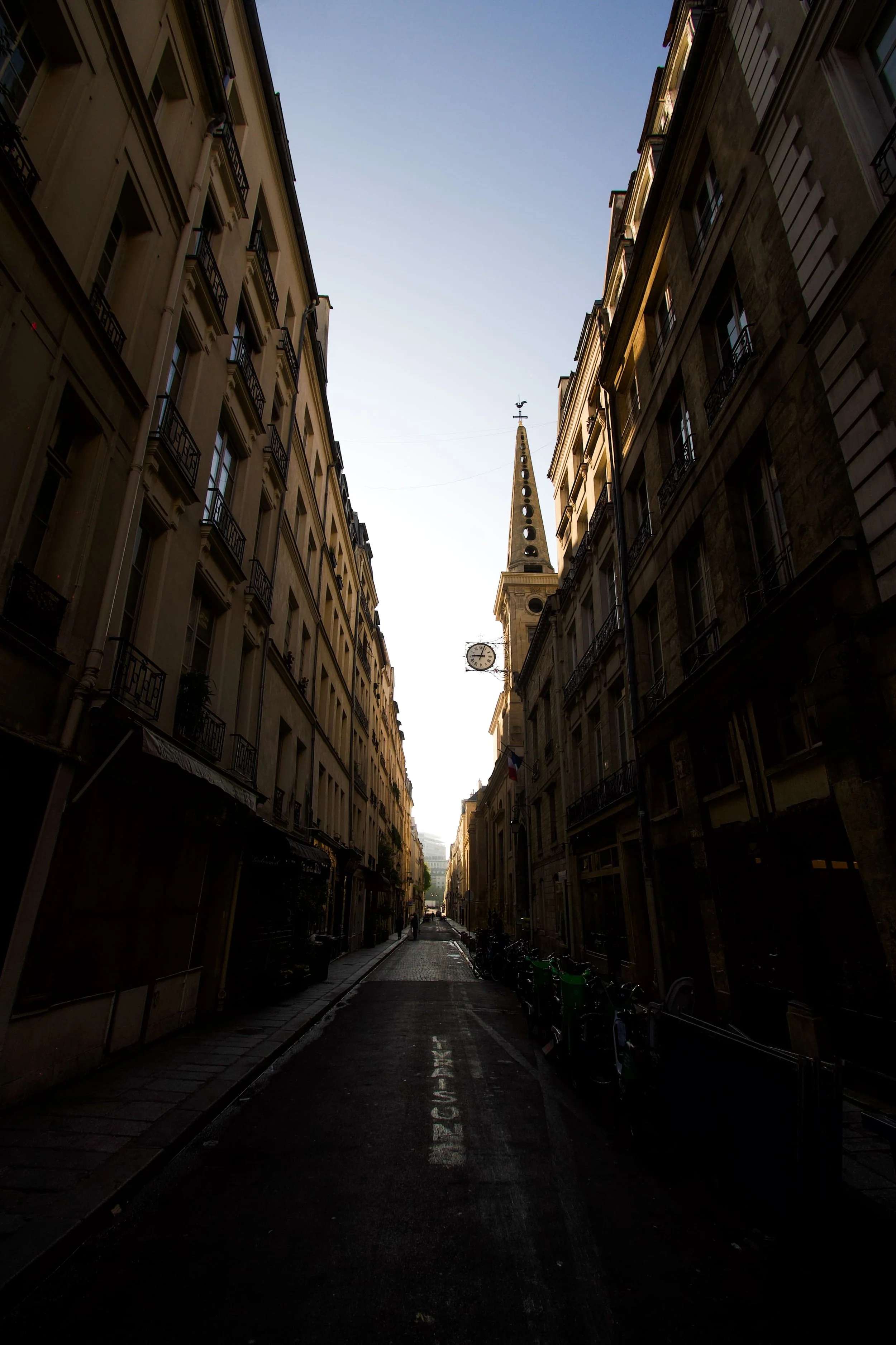 View of an urban alley lined with Haussmann buildings, with a church with its pointed steeple in the background, under a clear sky.